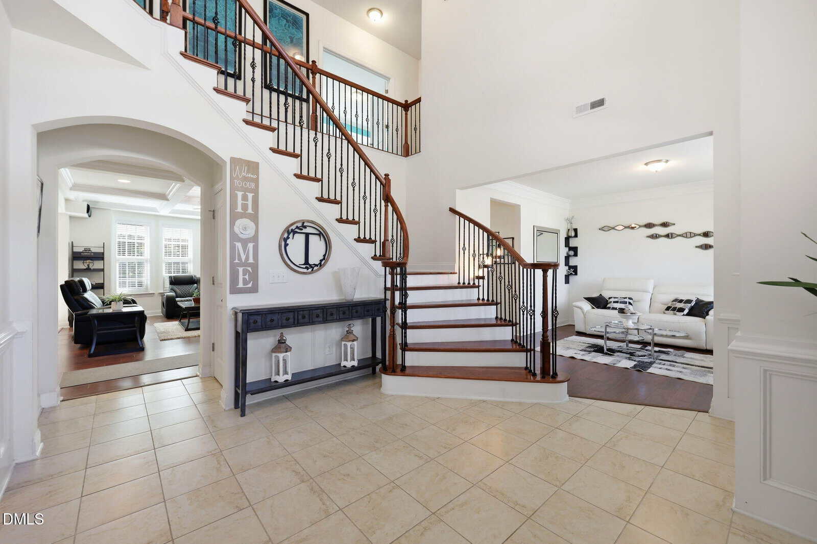Spacious foyer with tiled floor, wooden staircase, white walls, decorative console with mirror, two lanterns, and entryways leading to living rooms with sofas and natural light.