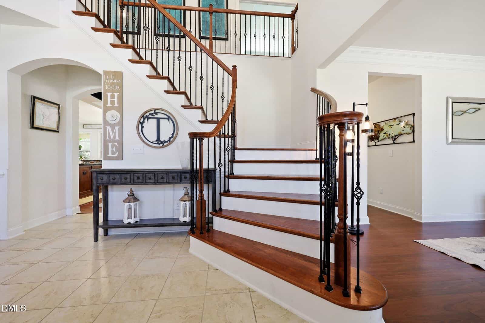 Curved wooden staircase with black iron rails in a bright entryway. Beige tile floor, white walls, and a console table with decor, including lanterns and a “HOME” sign on the wall. Doorways lead to other rooms.