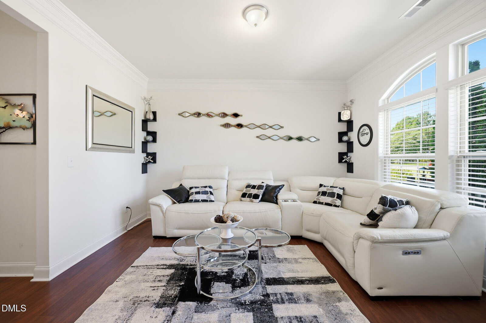 Modern living room with white sectional sofa, glass coffee table, black and white rug, wall art, and large windows letting in natural light. Decor includes throw pillows and a decorative bowl on the table.