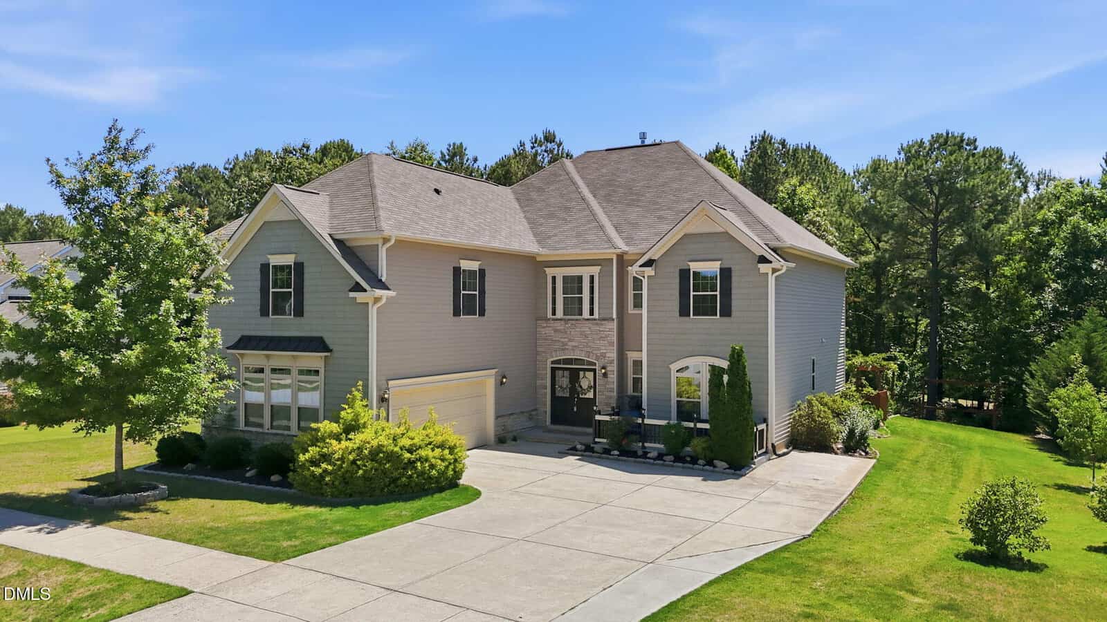 Two-story gray suburban house with a double garage, black front door, and manicured landscaping. A large driveway curves in front, and the house is surrounded by grass and trees under a clear blue sky.