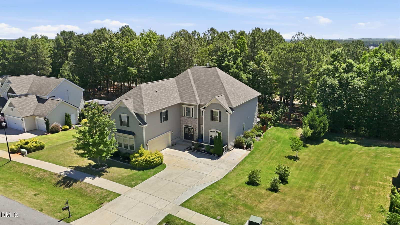 A two-story suburban house with a gray exterior, attached garage, and well-maintained front yard sits beside a spacious green lawn and driveway, bordered by trees and neighboring houses under a clear blue sky.