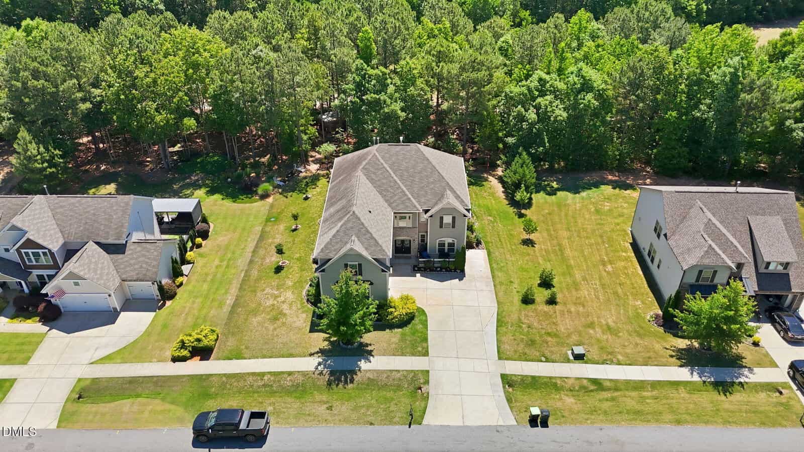 Aerial view of a two-story house with a large driveway and manicured lawn, flanked by similar neighboring homes. Trees line the backyard, and a car is parked on the street in front.