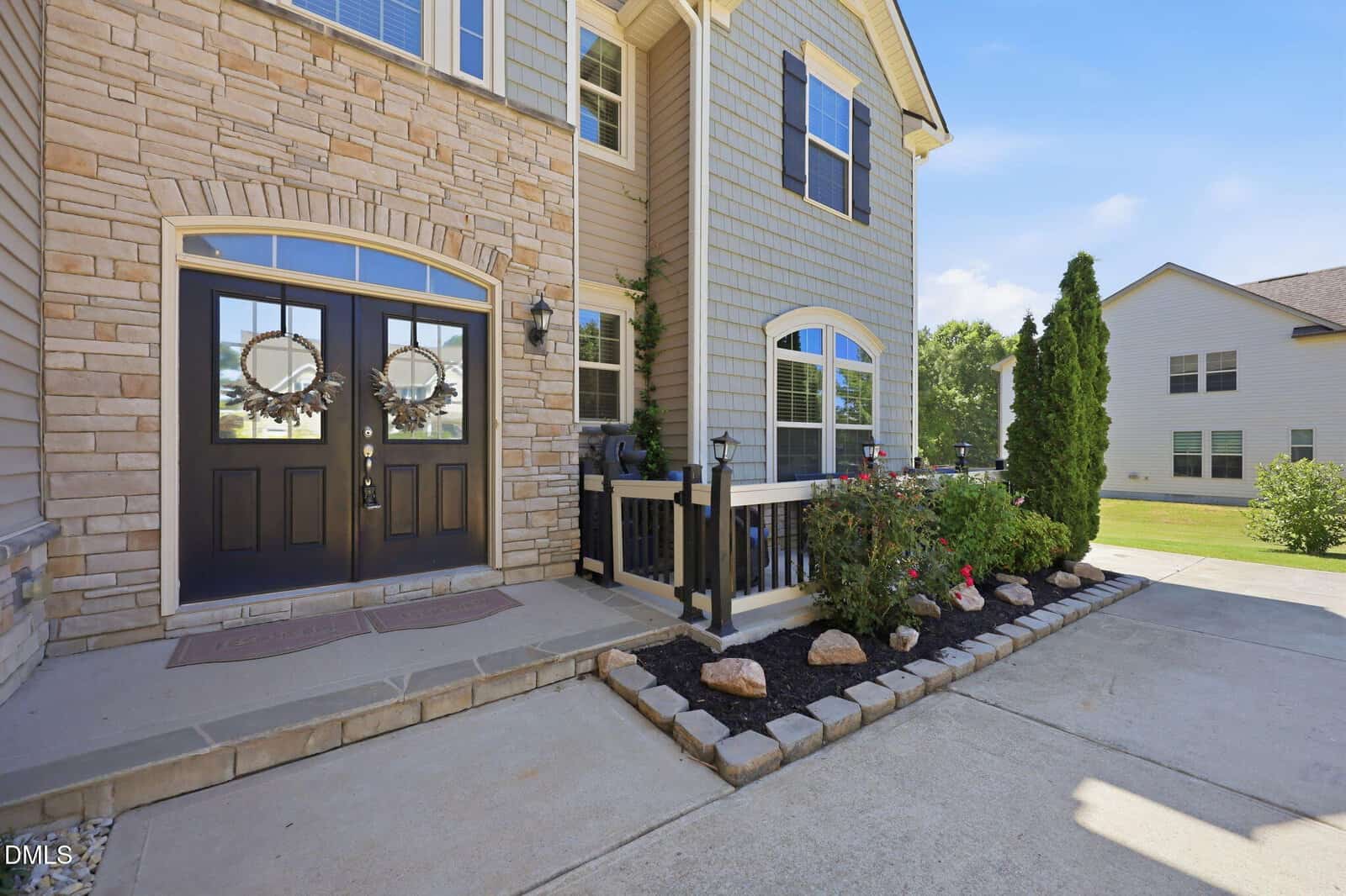 Front entrance of a modern house with double black doors, stone and siding exterior, a small porch with railing, landscaped flower bed with rocks, and a concrete driveway on a sunny day.