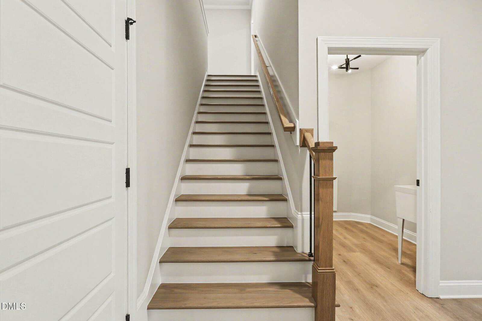 A modern, narrow staircase with wooden steps and a wooden handrail leads upward between white walls; an open doorway to the right reveals a room with light wood flooring and white trim.