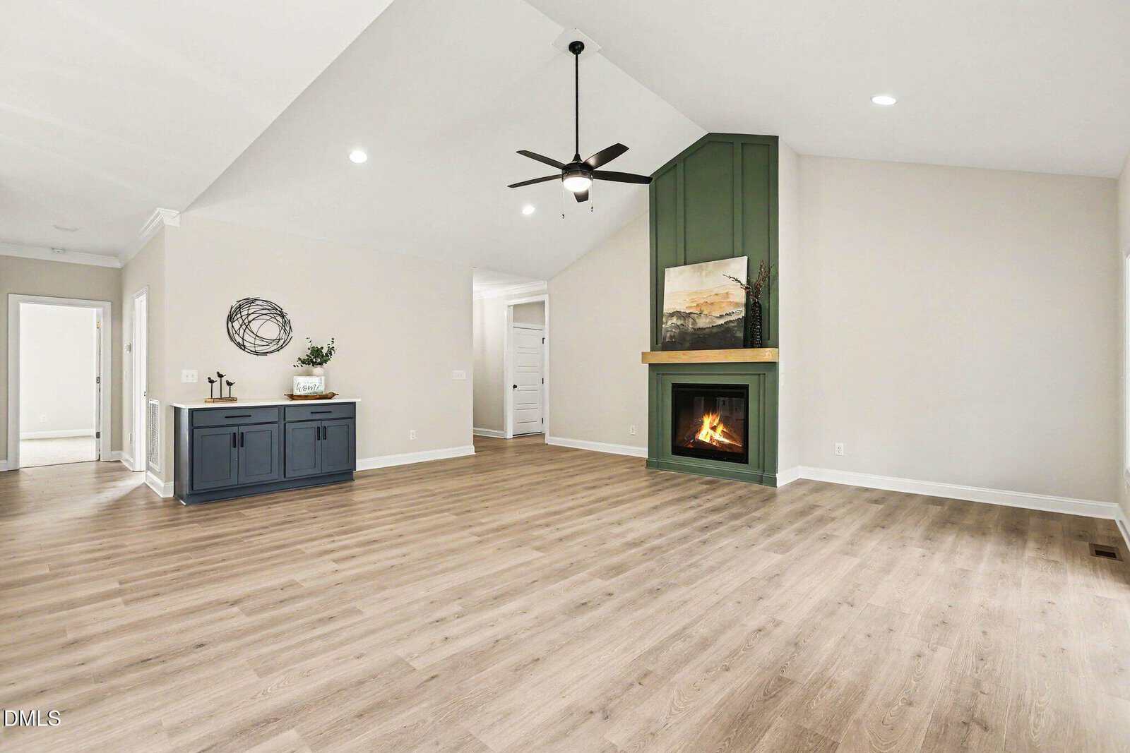 Spacious living room with light wood flooring, a green accent fireplace with a mantle and art, ceiling fan, recessed lighting, and a blue side cabinet against a white wall. Doorways lead to other rooms.