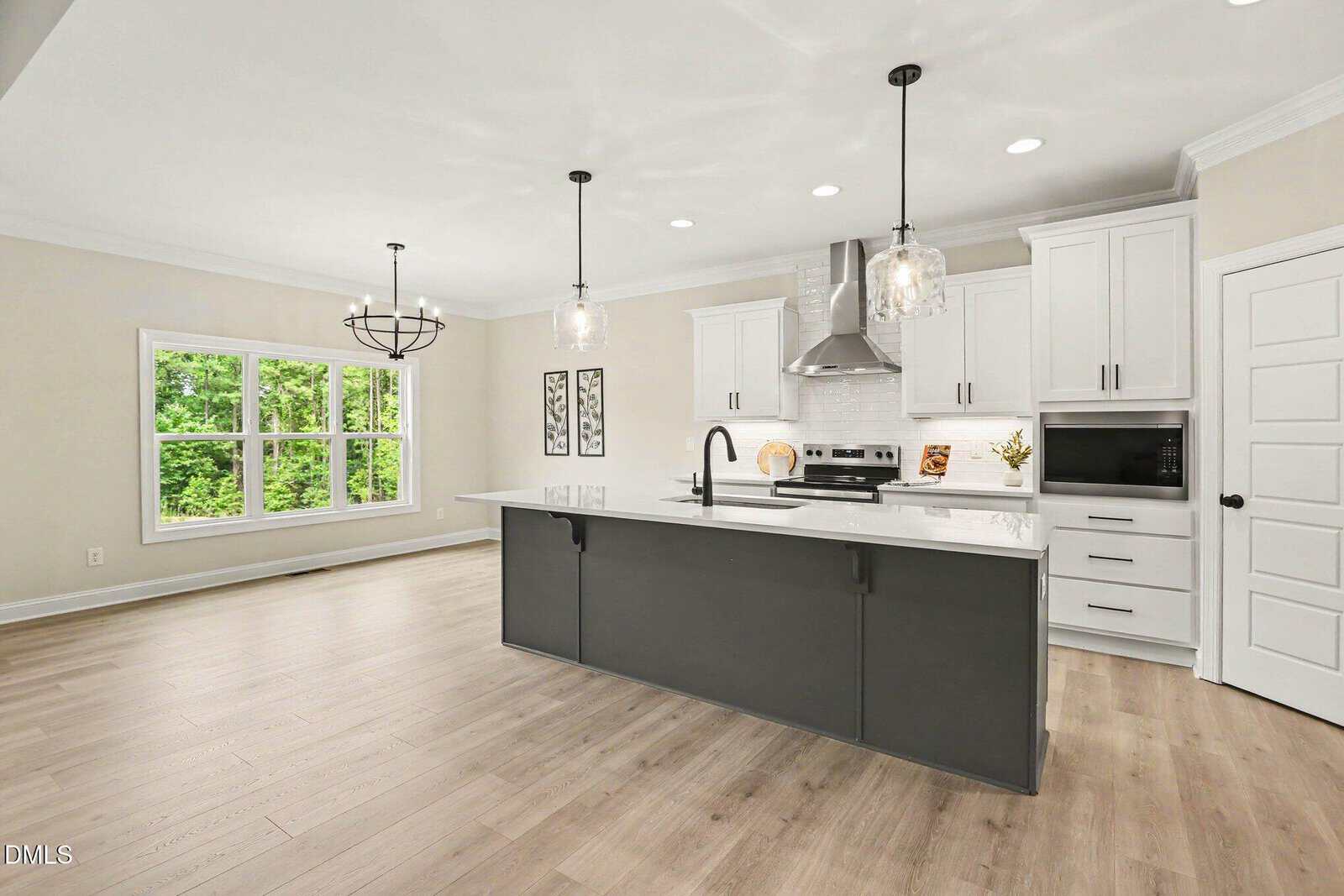 Modern kitchen with white cabinets, black island, pendant lights, stainless steel appliances, light wood floors, large windows, and a dining area with a chandelier. Neutral color palette with greenery visible outside.