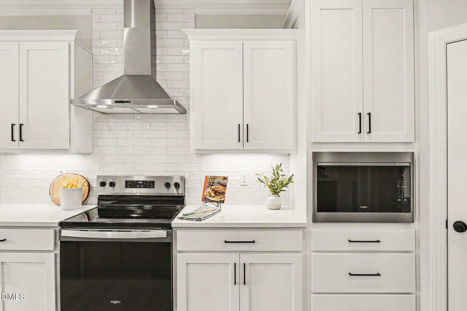 Modern kitchen with white cabinets, stainless steel stove and hood, microwave, subway tile backsplash, a small plant, a cookbook, and utensils in a white container on the countertop.