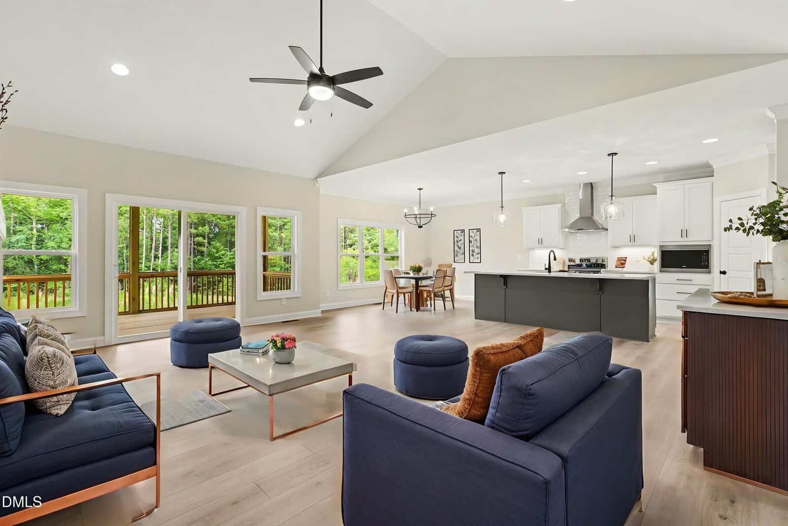Spacious open-concept living room and kitchen with navy sofas, light wood floors, large windows, modern lighting, a center island, and views of a green backyard through multiple glass doors.