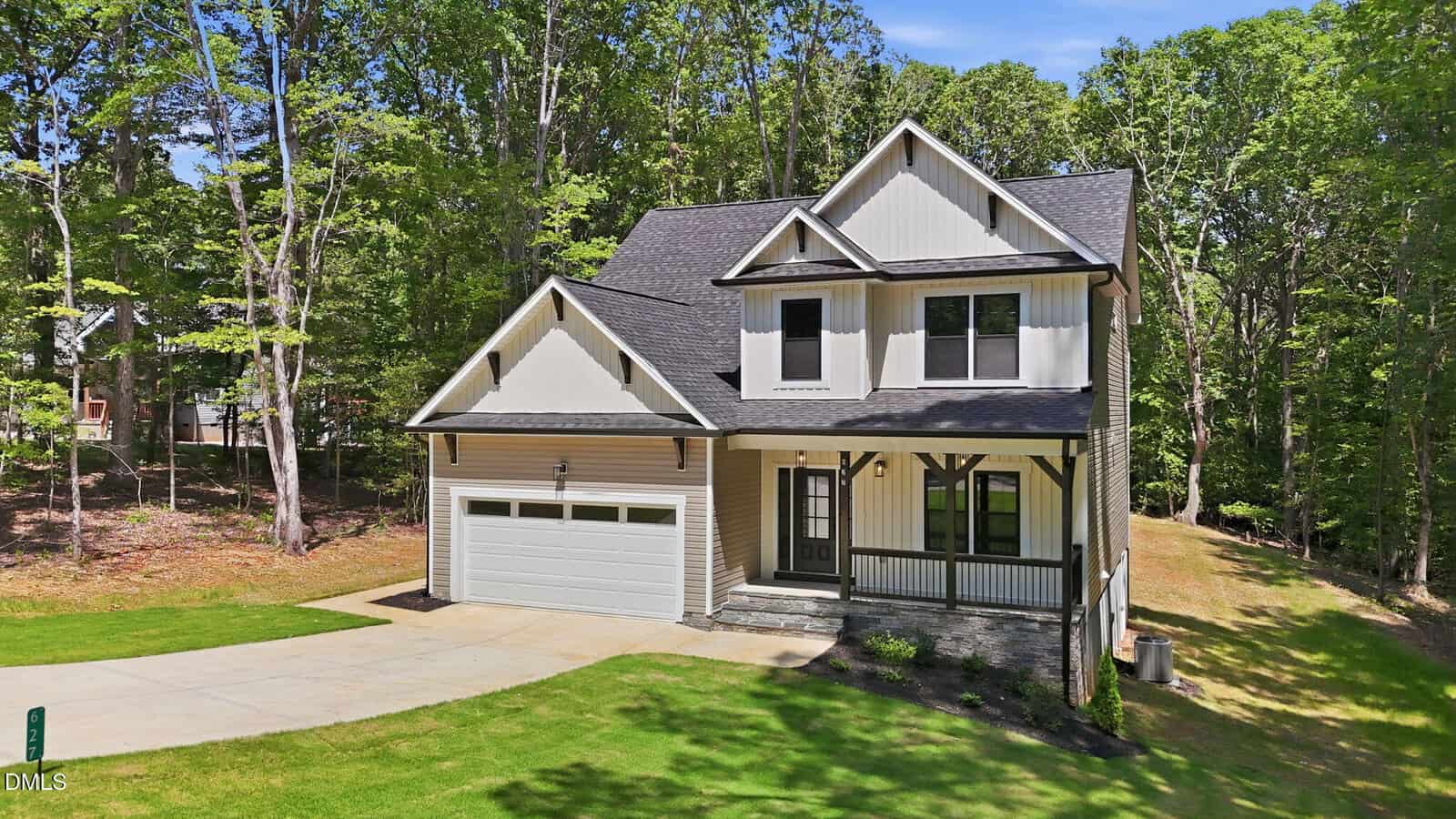 A two-story modern house with light-colored siding, dark trim, a front porch, and an attached two-car garage. The home sits on a green lawn, surrounded by trees under a blue sky.