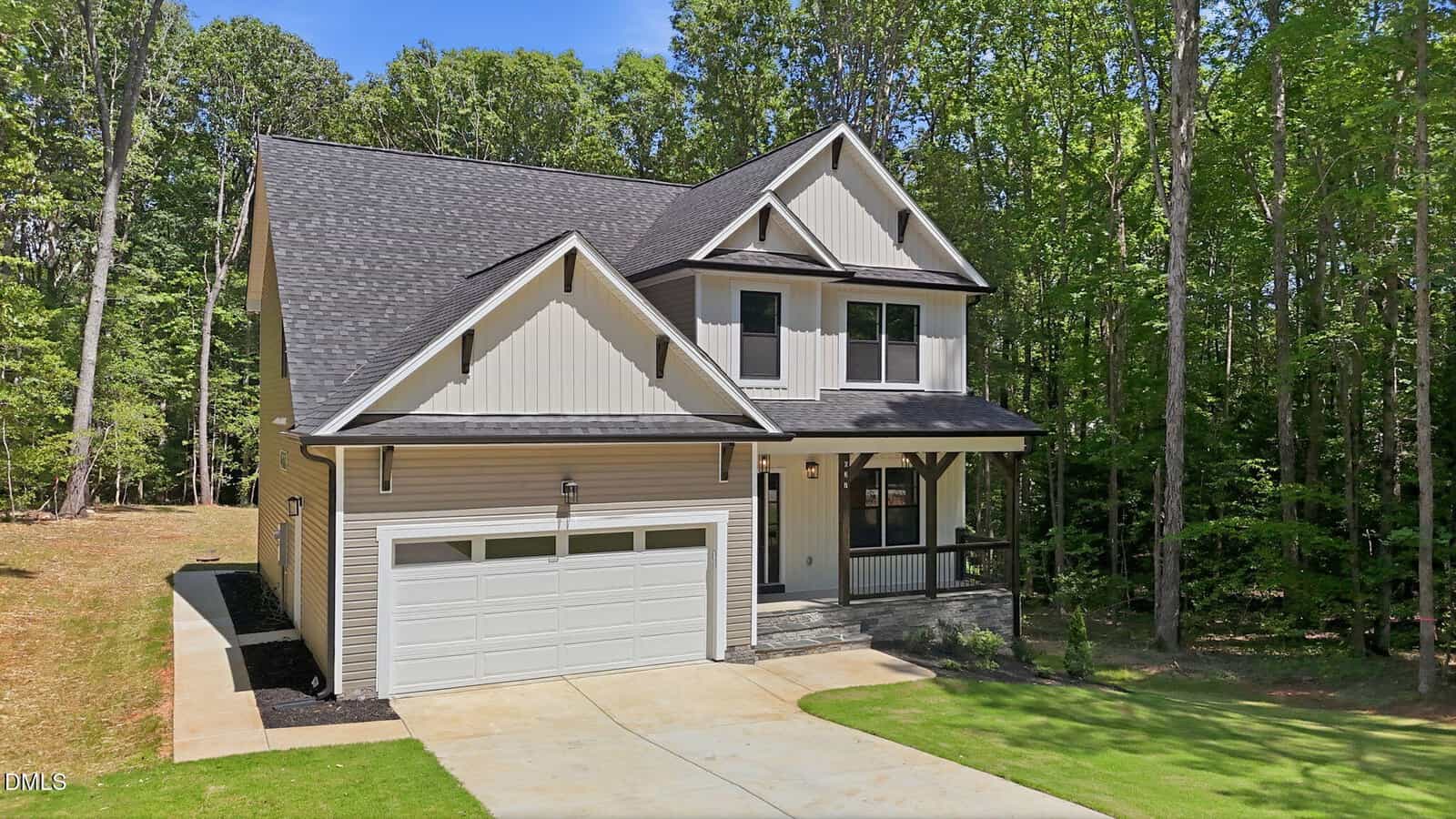 Two-story modern house with light-colored siding, dark trim, and a covered front porch, surrounded by tall trees and greenery. The home features a double garage and a paved driveway.