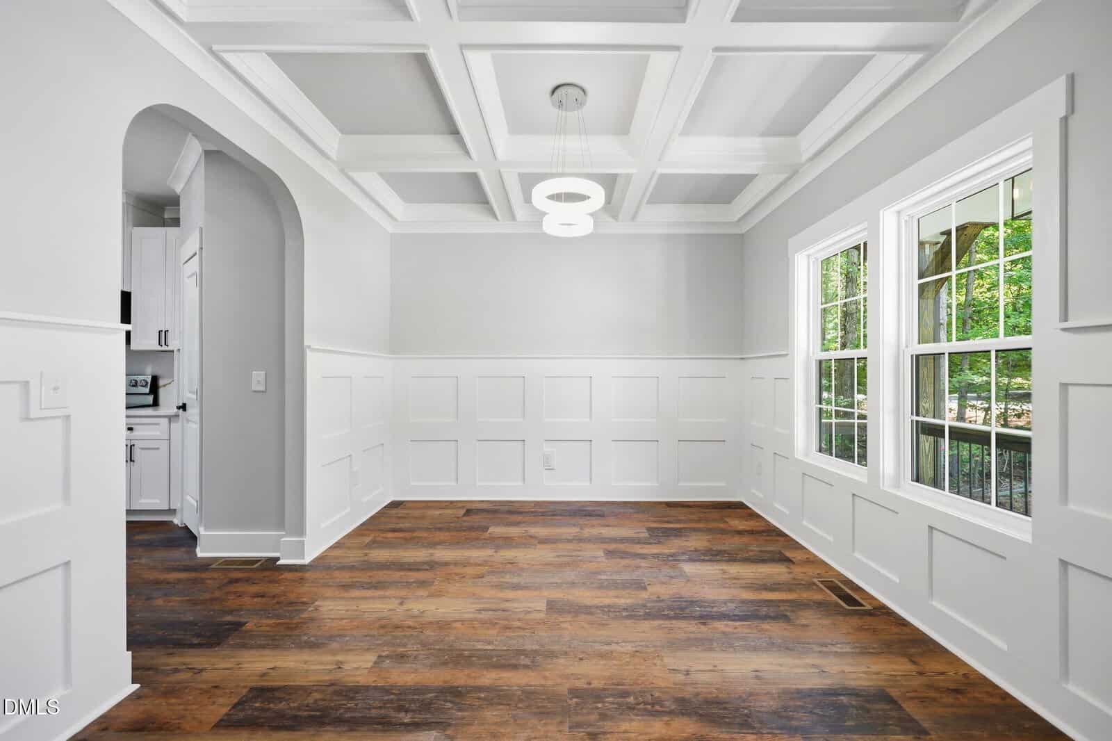 A bright dining room with white wainscoting, coffered ceiling, modern pendant light, large windows, and wood-look flooring. An arched doorway leads to a kitchen with white cabinetry.