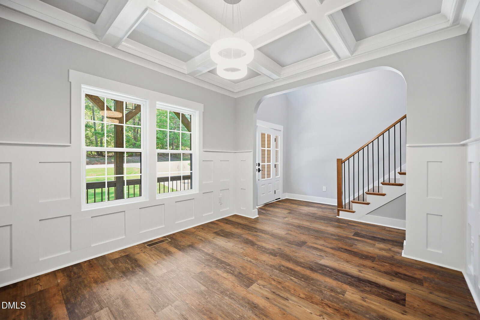 Bright room with coffered ceiling, white walls, and wood floors. Large window on the left lets in natural light. An archway leads to a staircase with a wooden handrail and black balusters near the entry door.