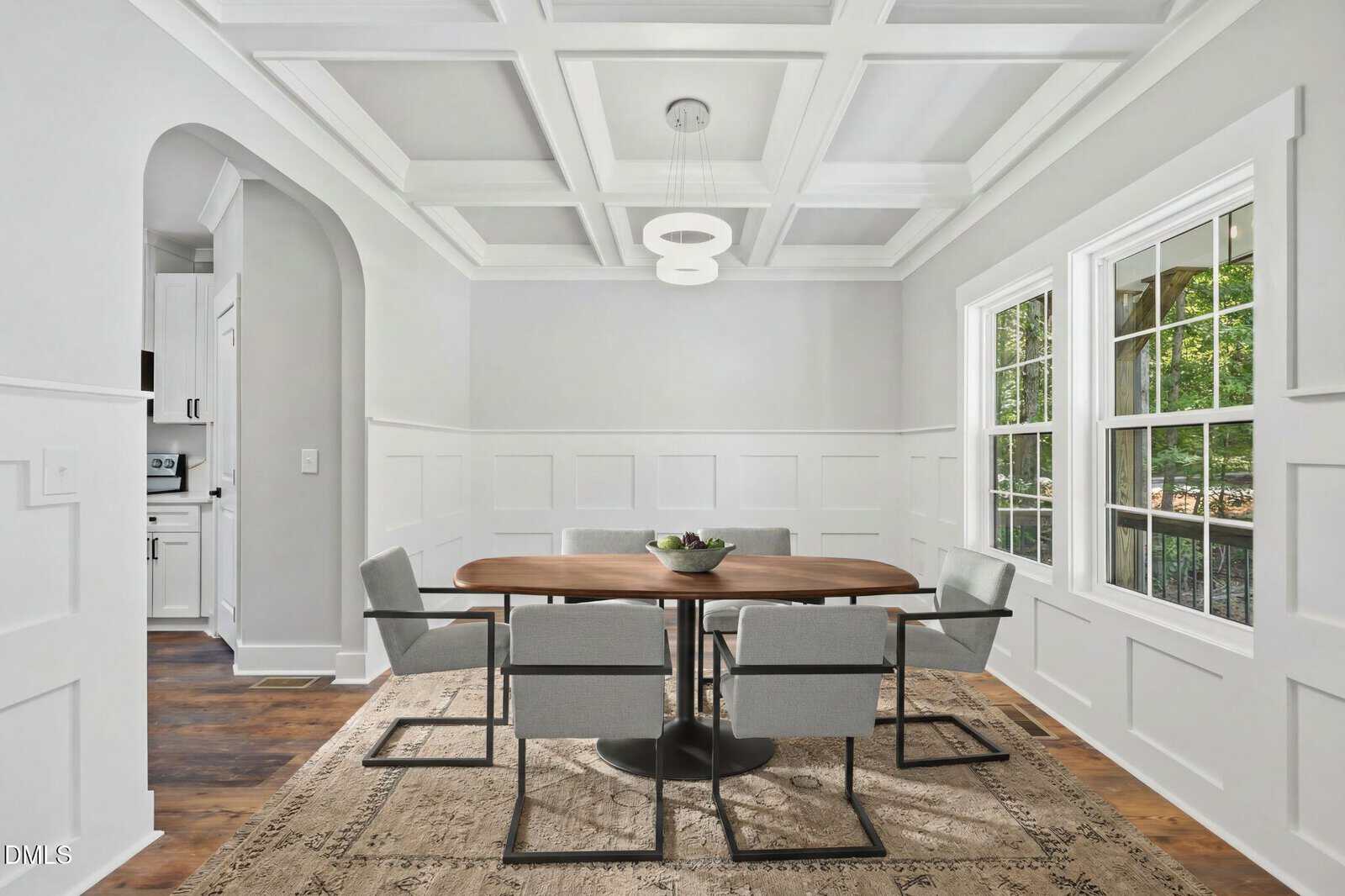 A modern dining room with white wainscoting, coffered ceiling, wooden table, six gray chairs, large windows, and a neutral area rug on a hardwood floor. A pendant light hangs above the table.