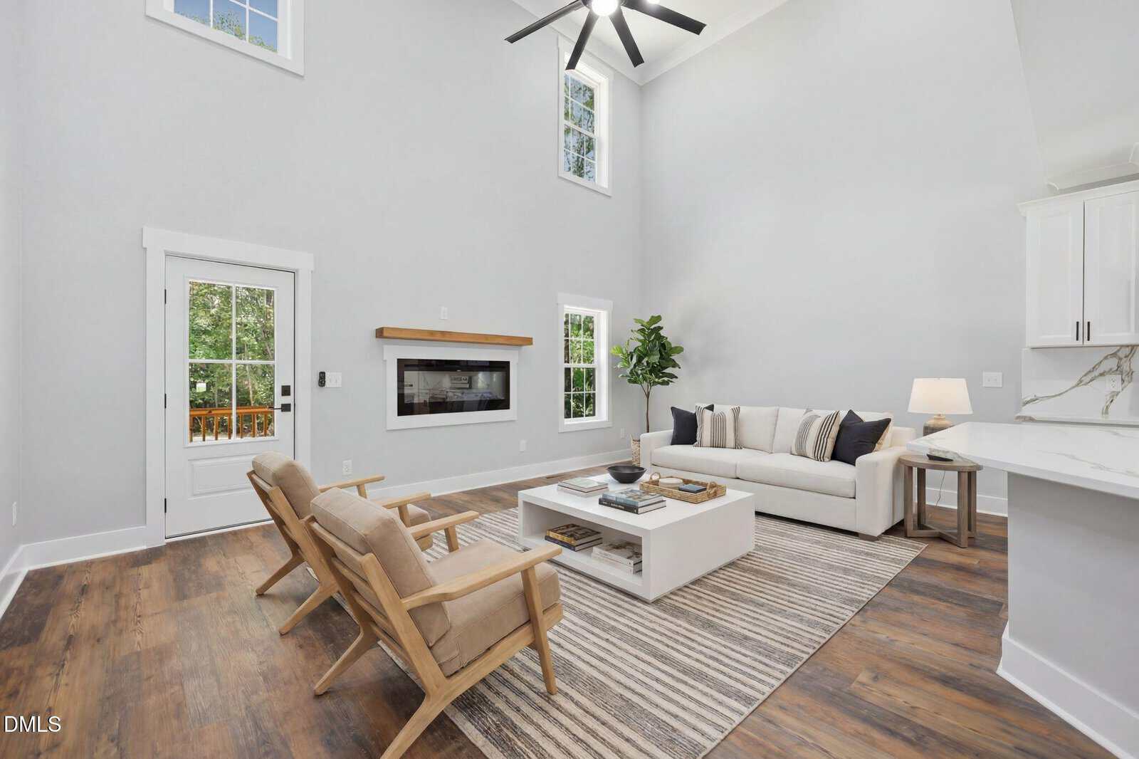 Bright living room with high ceilings, modern decor, white sofa, two wooden armchairs, striped rug, wall-mounted fireplace, large windows, and a glass door leading outside. Green plant and neutral tones create a cozy atmosphere.