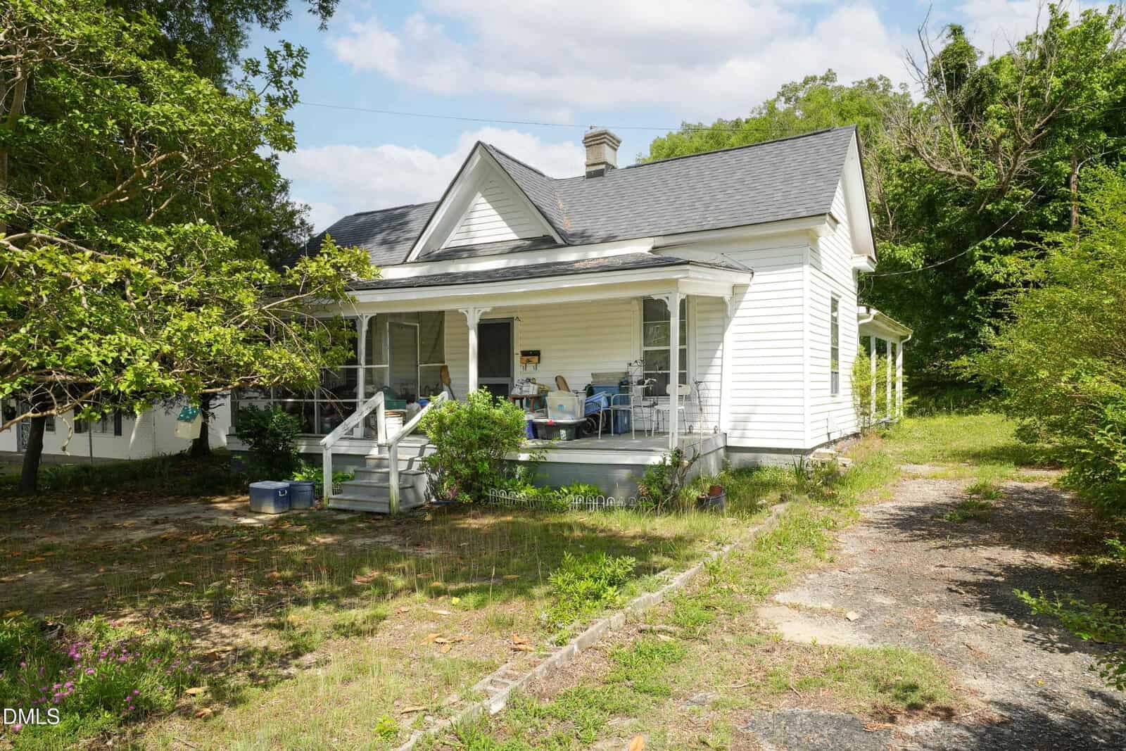 A charming white, single-story house at 604 S Main Street in Louisburg with a covered front porch, several chairs, and surrounded by trees and greenery. The yard features patches of grass and a stone-lined walkway leading to the porch.
