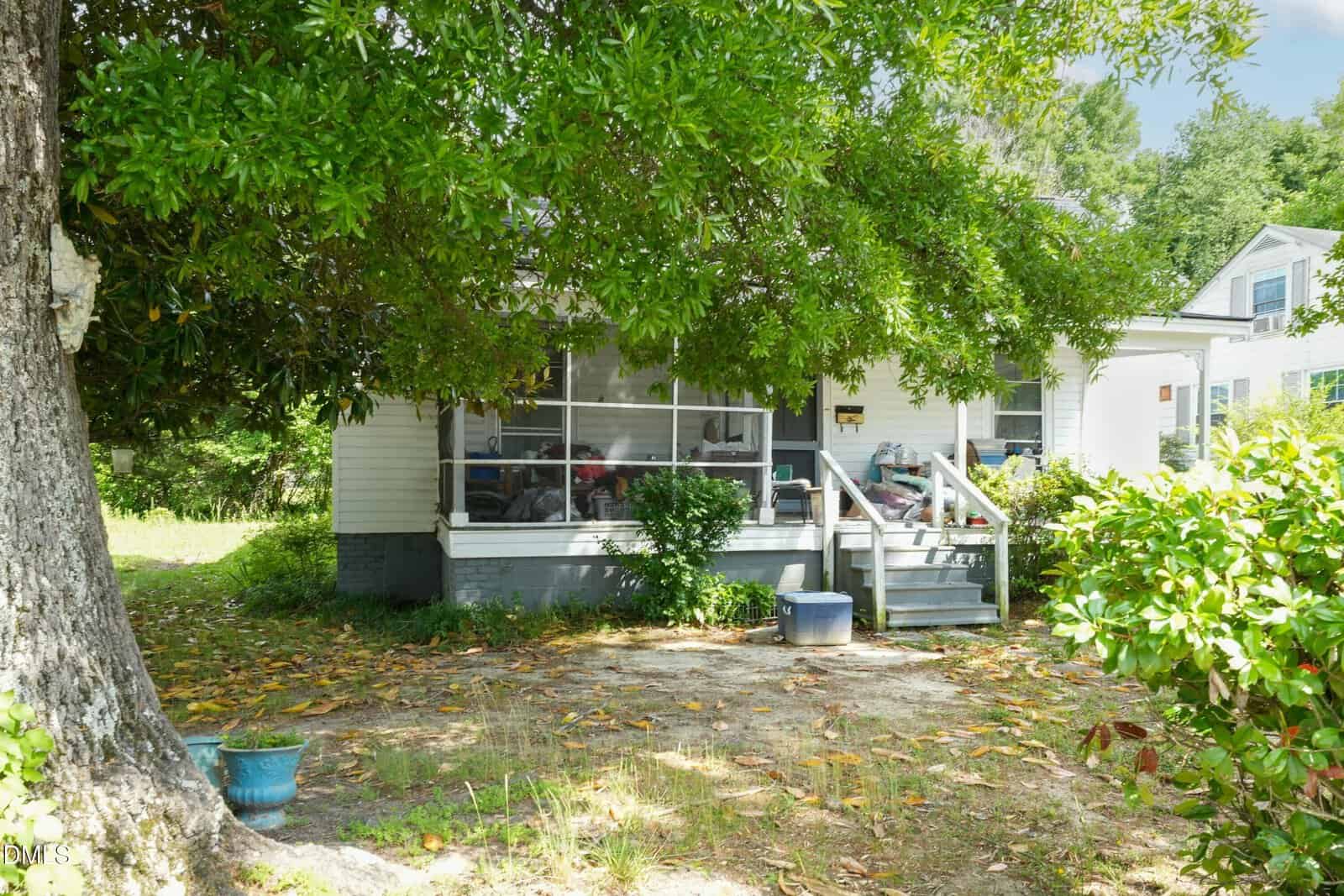 A small white house with a screened-in porch, front steps, and surrounded by leafy trees and bushes. Fallen leaves cover the yard, and potted plants sit near the steps under the shade of a large tree.