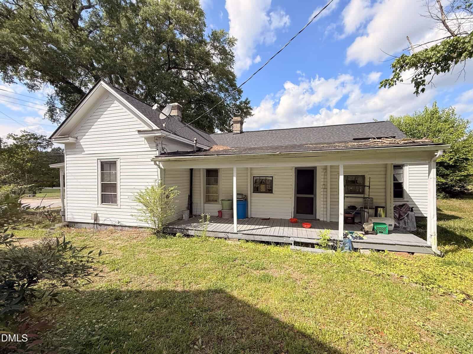 A white, weathered house with a covered front porch, some peeling paint, and a few items scattered on the porch; set against a grassy yard and blue sky with clouds.