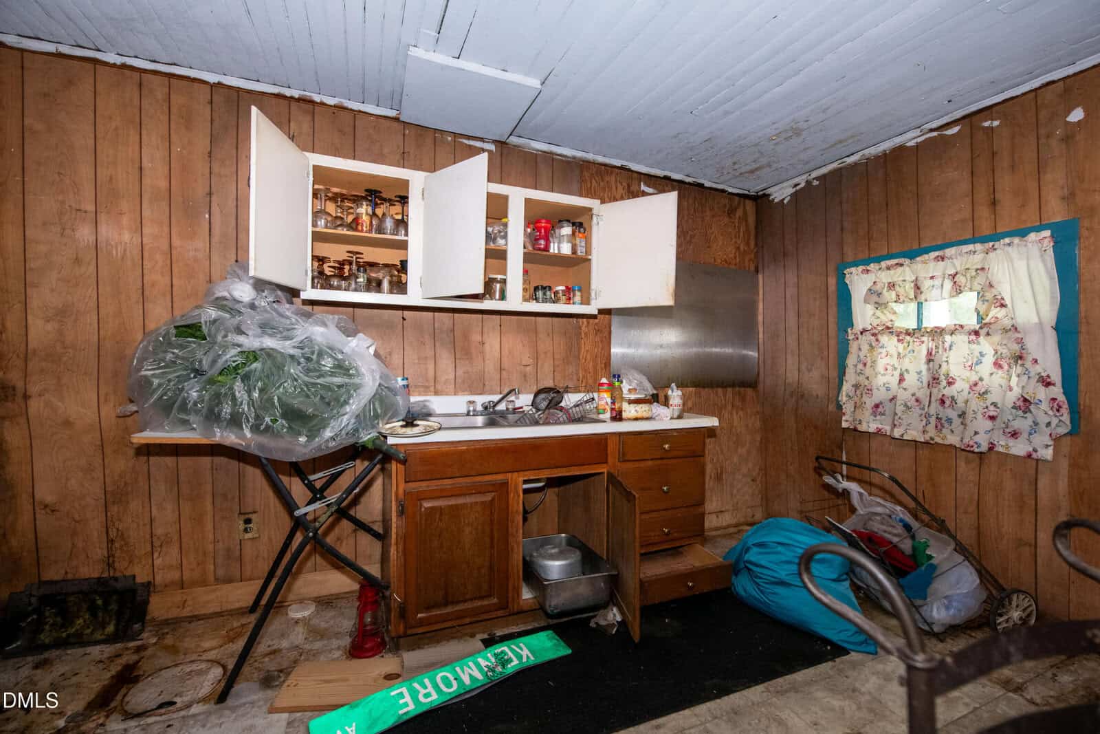 A cluttered kitchen with wood-paneled walls, open cabinets revealing jars and containers, a messy countertop, cleaning supplies, plastic bags, a laundry basket, and a window with floral curtains.