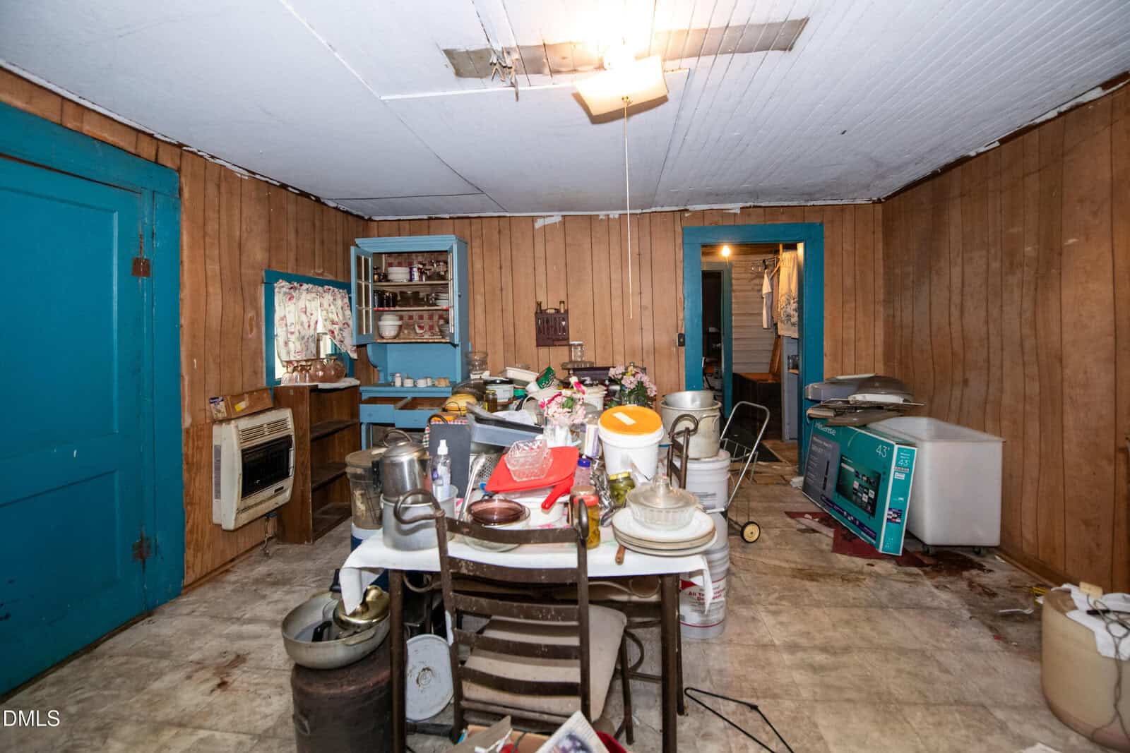 A cluttered, outdated room with wood-paneled walls, a table piled with kitchen items, scattered chairs, and bright blue door frames. Appliances and household goods are strewn across the worn floor. Ceiling tiles appear damaged.