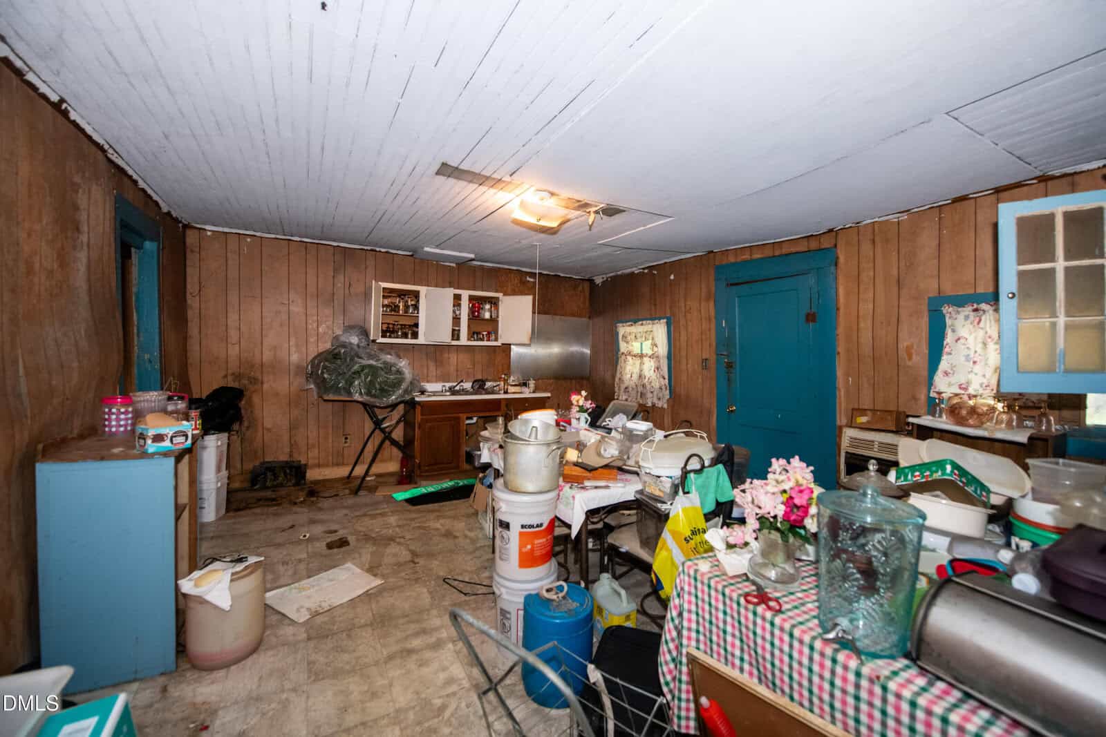 A cluttered kitchen with wood-paneled walls, blue doors, a checkered tablecloth, various appliances, and household items scattered across tables and counters. The ceiling and floor show signs of wear.
