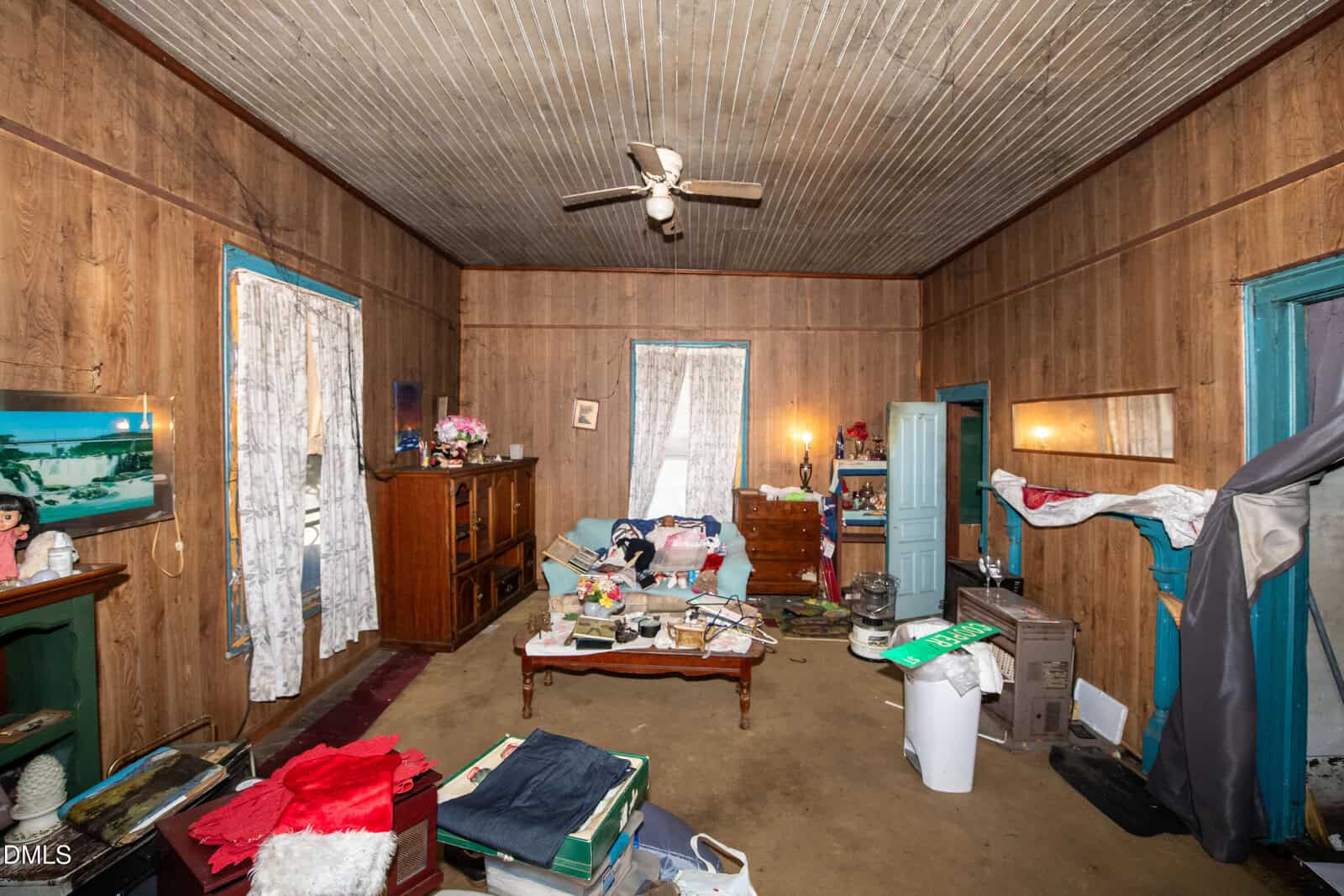 Cluttered living room with wood-paneled walls, a ceiling fan, two windows with lace curtains, various furniture, boxes, and items scattered on the floor. Light comes from lamps and window.