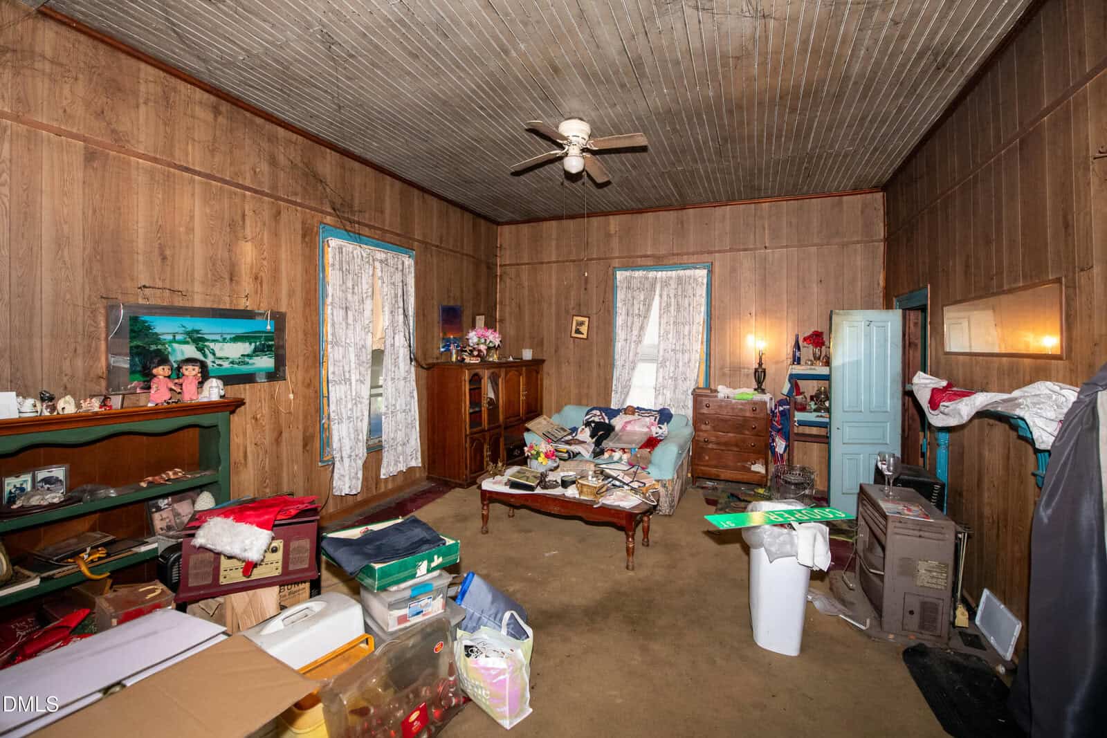 A cluttered, rustic bedroom with wood panel walls, a single bed, boxes, scattered belongings, a green mantle, two windows with lace curtains, a ceiling fan, and various pieces of vintage furniture.