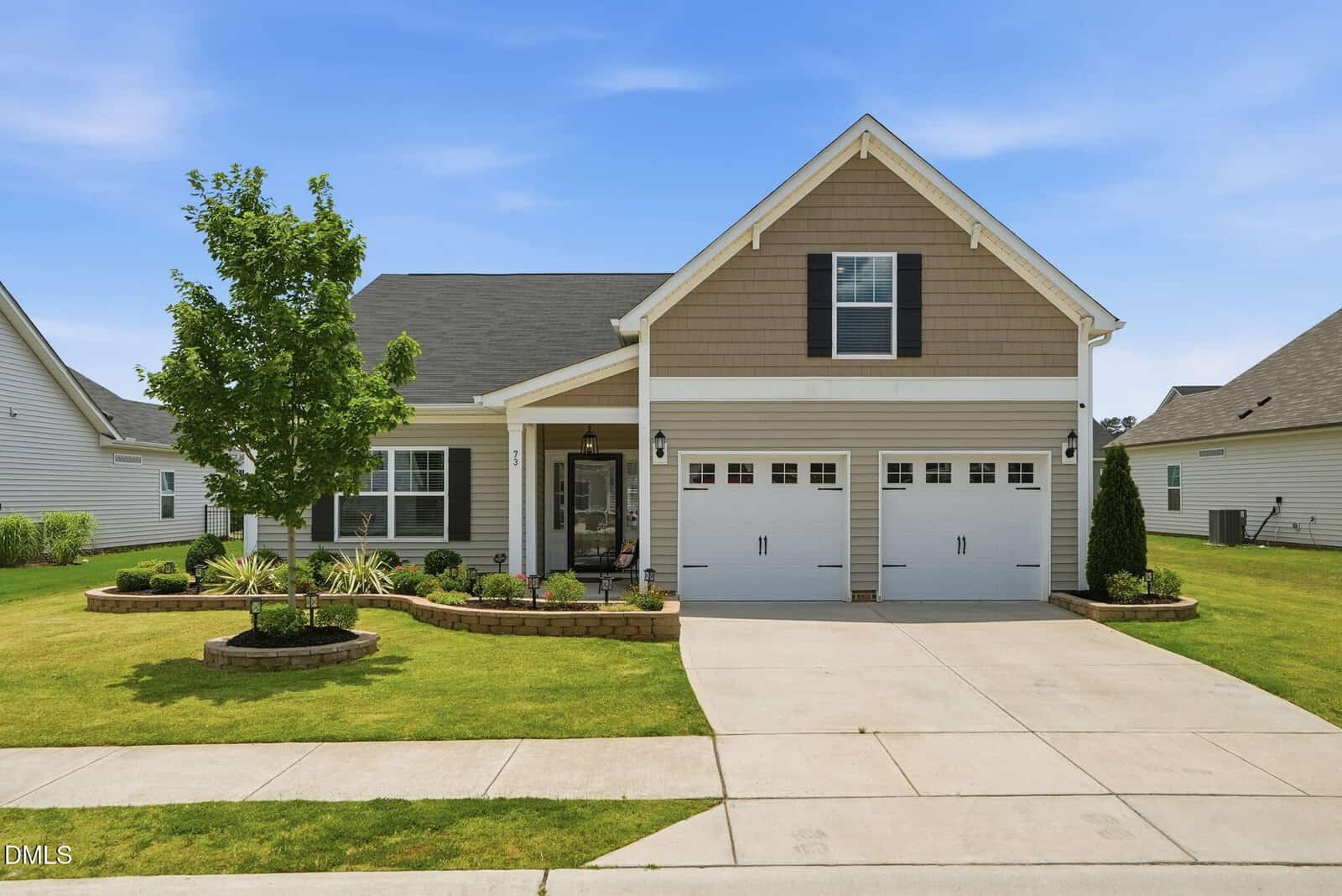 A modern single-story house with beige siding, a double garage, manicured lawn, a young tree, and landscaped shrubs under a clear blue sky.