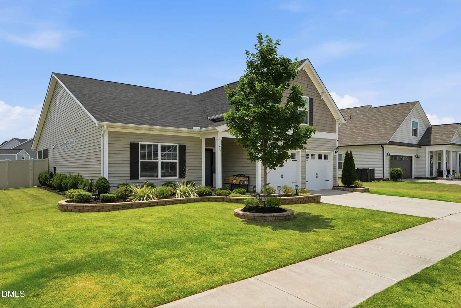 Single-story house with gray siding, dark shutters, and a gable roof. The front yard features neatly trimmed grass, landscaped plants, a small tree, and a driveway leading to a white garage. Bright, clear sky overhead.