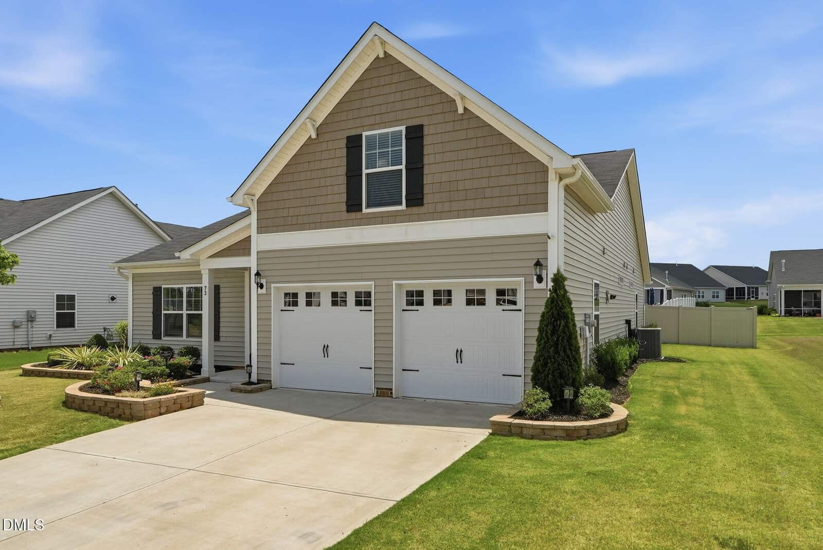 Single-story suburban house with tan siding, white trim, and a double garage. Manicured lawn, landscaped flowerbeds, and a clear blue sky in the background. Neighboring houses are visible on both sides.