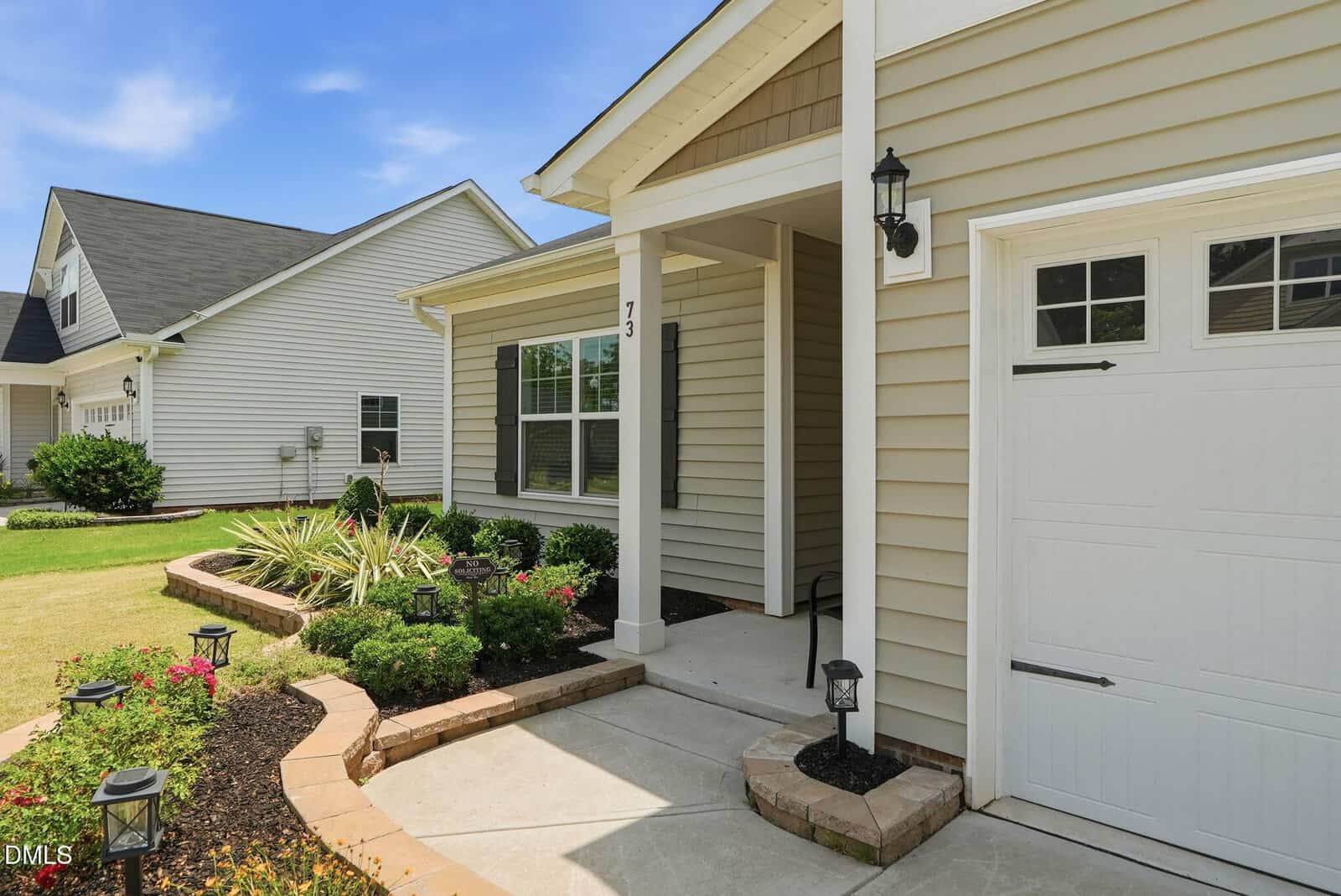 Single-story beige house with white trim, a small covered porch, and a white garage door. There are manicured shrubs, flowers, and pathway lights along a curved stone-edged walkway in the front yard.