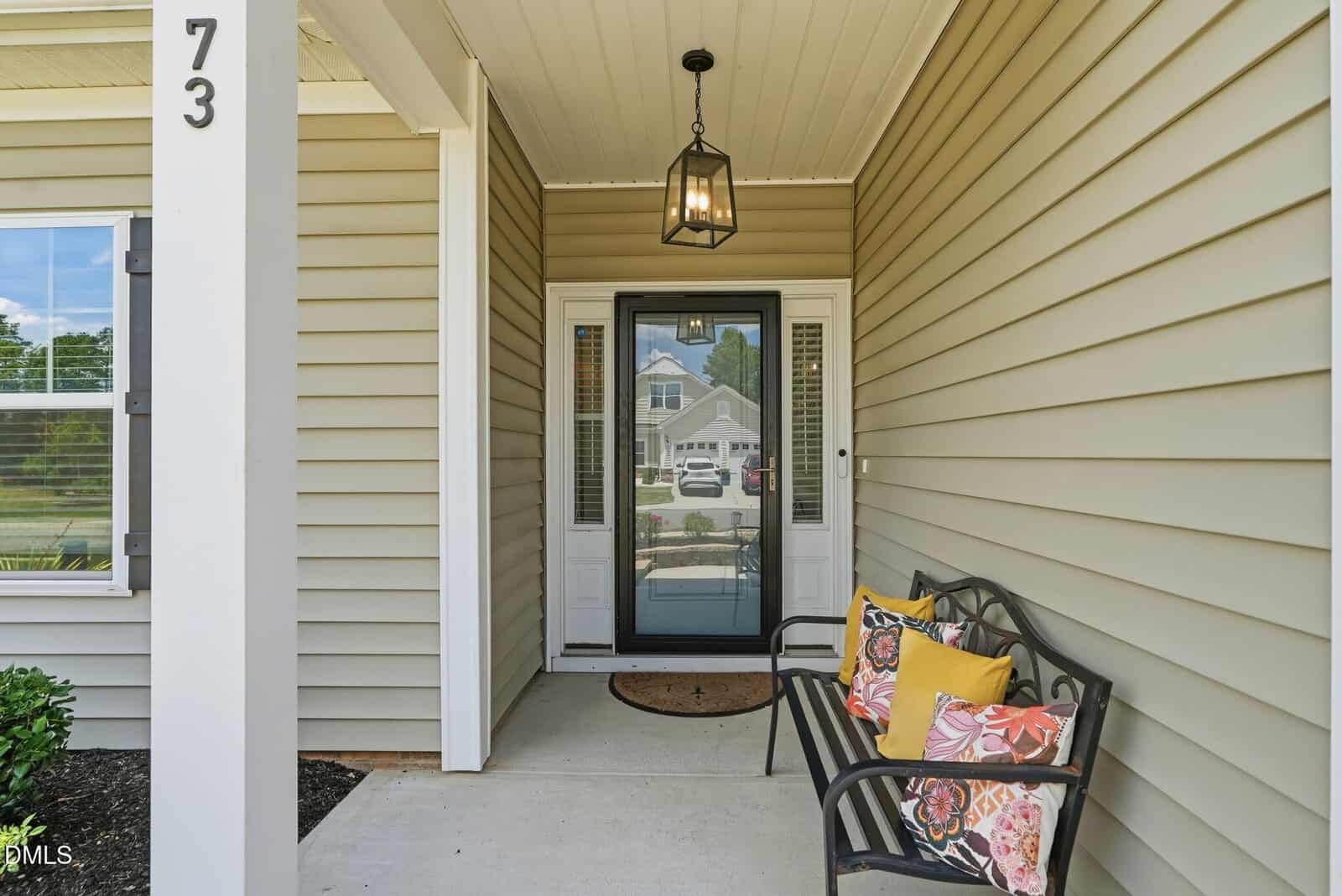 A covered front porch with beige siding, a black bench with colorful pillows, a glass front door with side windows, lantern light fixture, house number 73, and a view of a street with cars and houses reflected in the door.