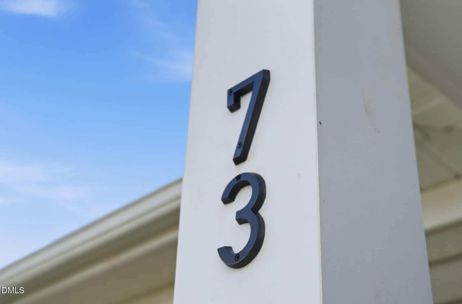 Black numbers “7” and “3” mounted vertically on a white post, with a blue sky and part of a roof visible in the background.