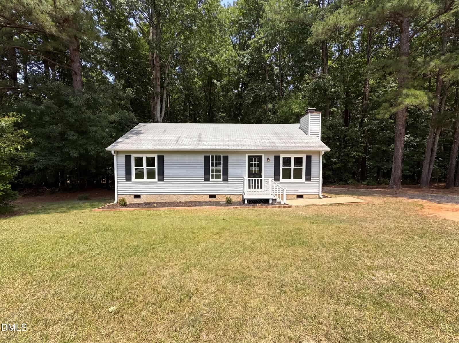 A small, single-story white house with a gray roof sits at 128 Mill Creek Drive in Youngsville, nestled before a wooded area. The front porch has steps and railing, and the grassy yard features a dirt path to the right.