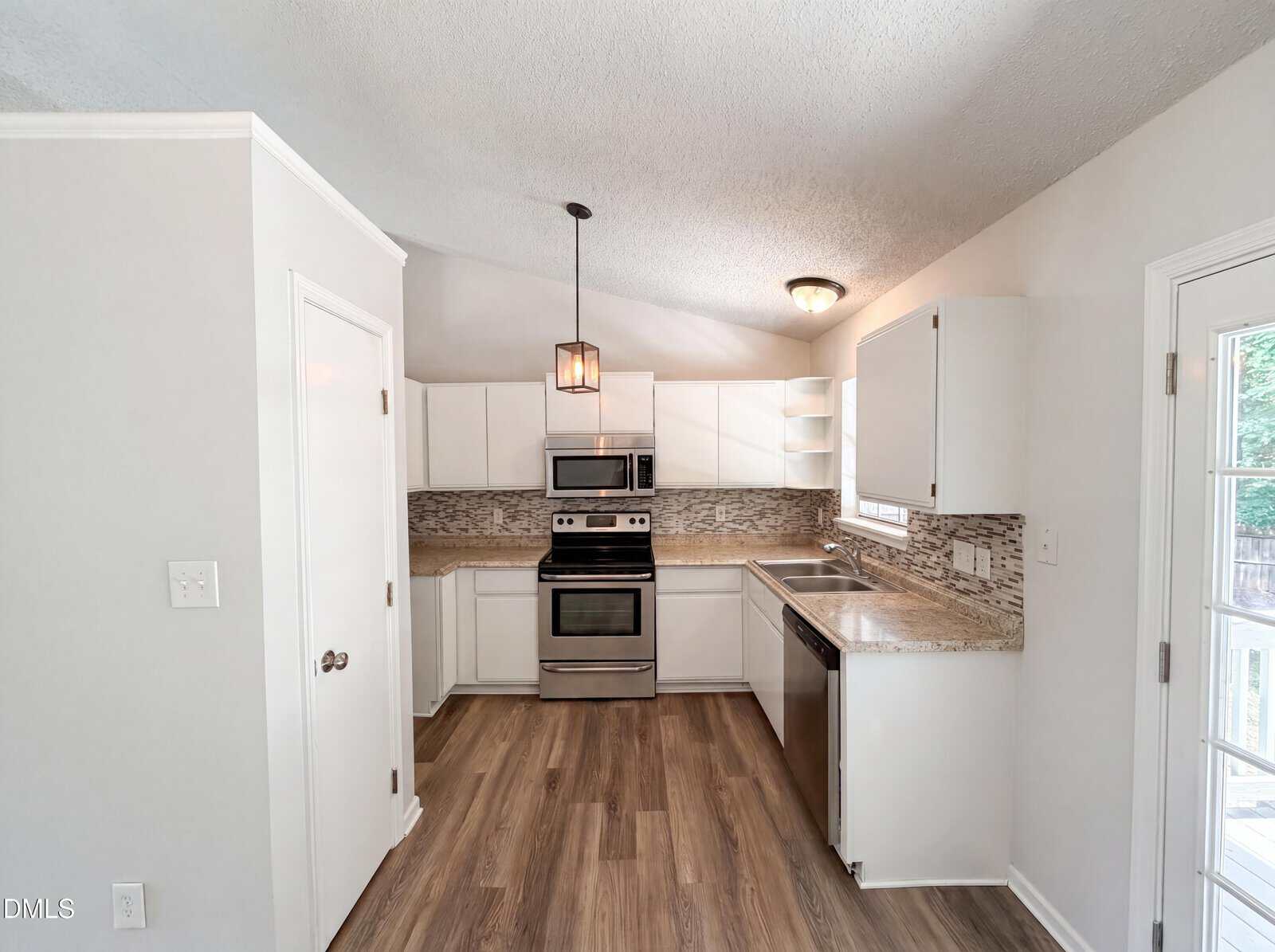 Modern kitchen with white cabinets, stainless steel appliances, granite countertops, a mosaic tile backsplash, wood-style flooring, and a pendant light above the stove. A door with glass panes is on the right.