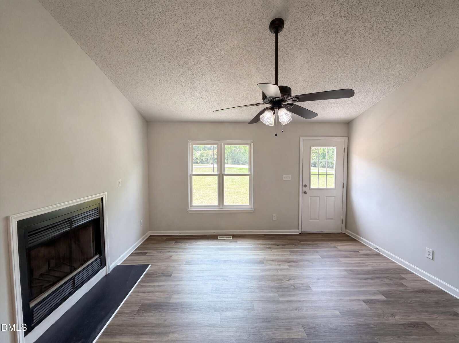 A bright, empty living room with light gray walls, wood flooring, a ceiling fan with lights, a fireplace on the left, and two windows next to a white door leading outside.