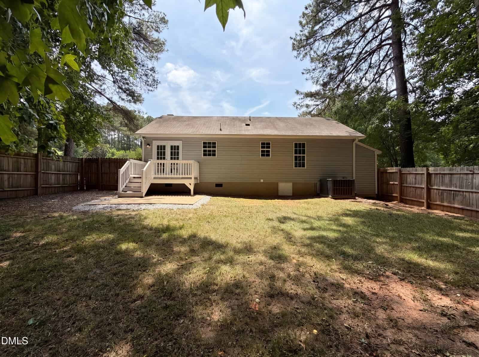 A fenced backyard with green grass and trees, featuring a beige single-story house. There is a small white deck with stairs leading to a patio area. The sky is partly cloudy.