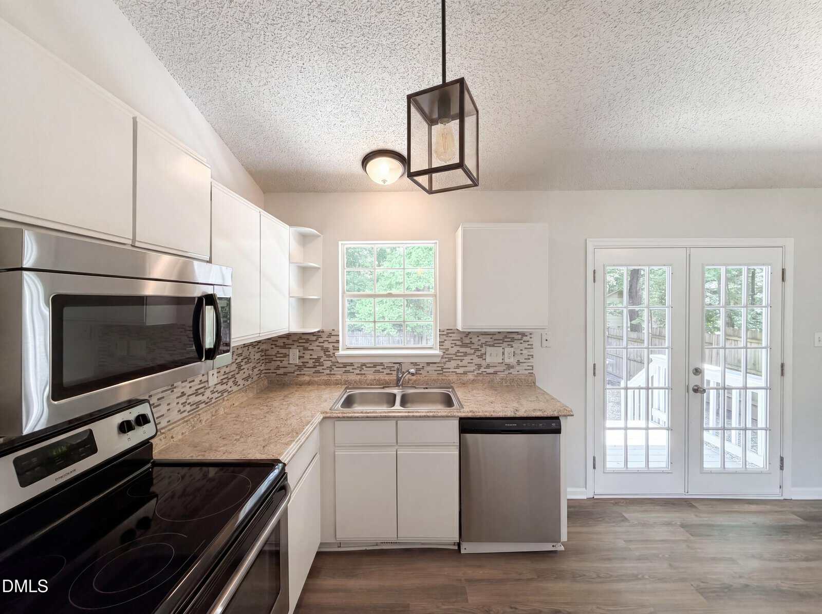 Modern kitchen with white cabinets, stainless steel appliances, double sink, tile backsplash, and wooden floors. Two windows and glass double doors let in natural light.