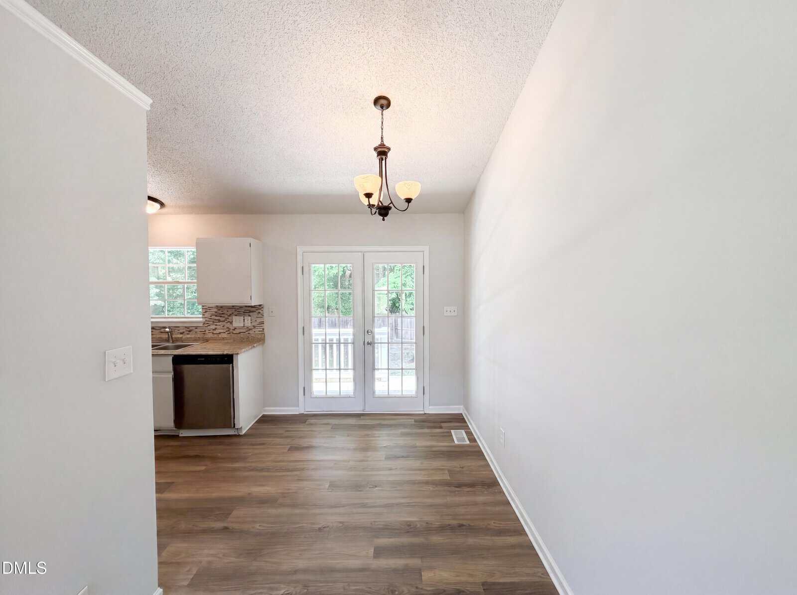 Bright dining area with wood flooring, chandelier, and French doors leading to a deck. Kitchen with white cabinets and stainless appliances is partially visible on the left. Light gray walls throughout.