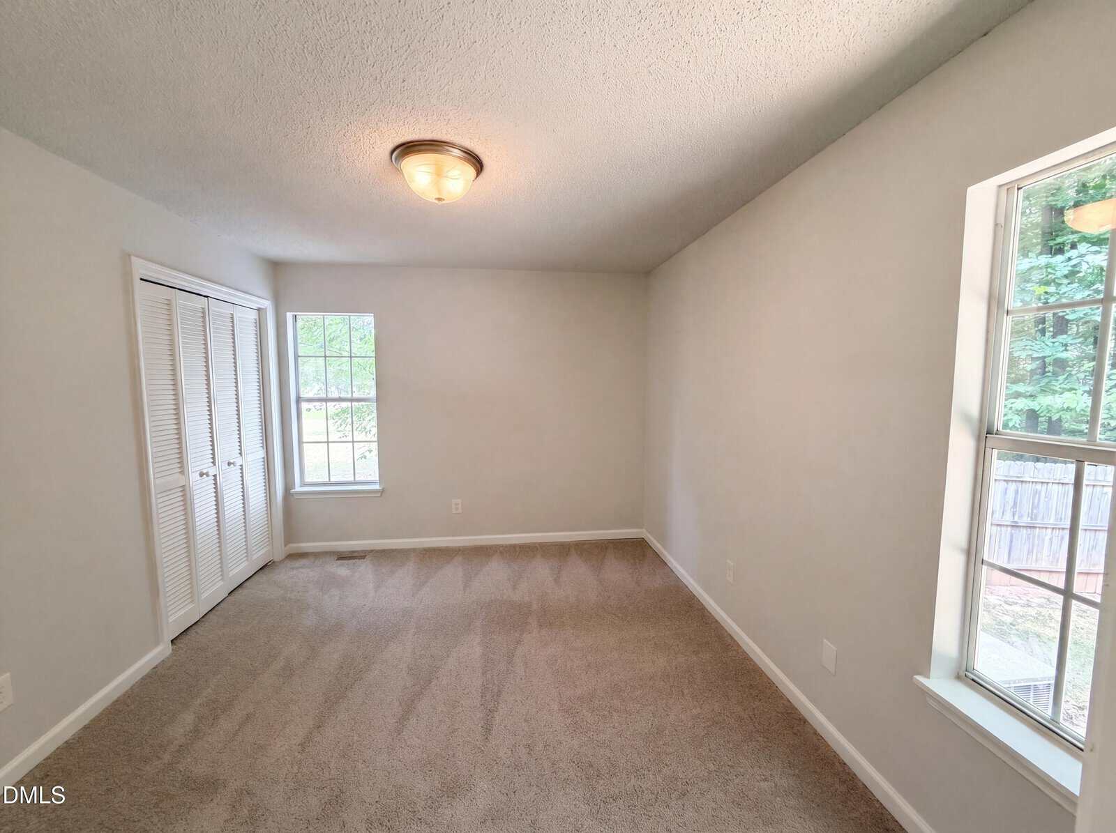 A small, empty room with beige carpet, white walls, two windows, a ceiling light, and a closed white closet door. Sunlight comes through the windows, highlighting the clean carpet.