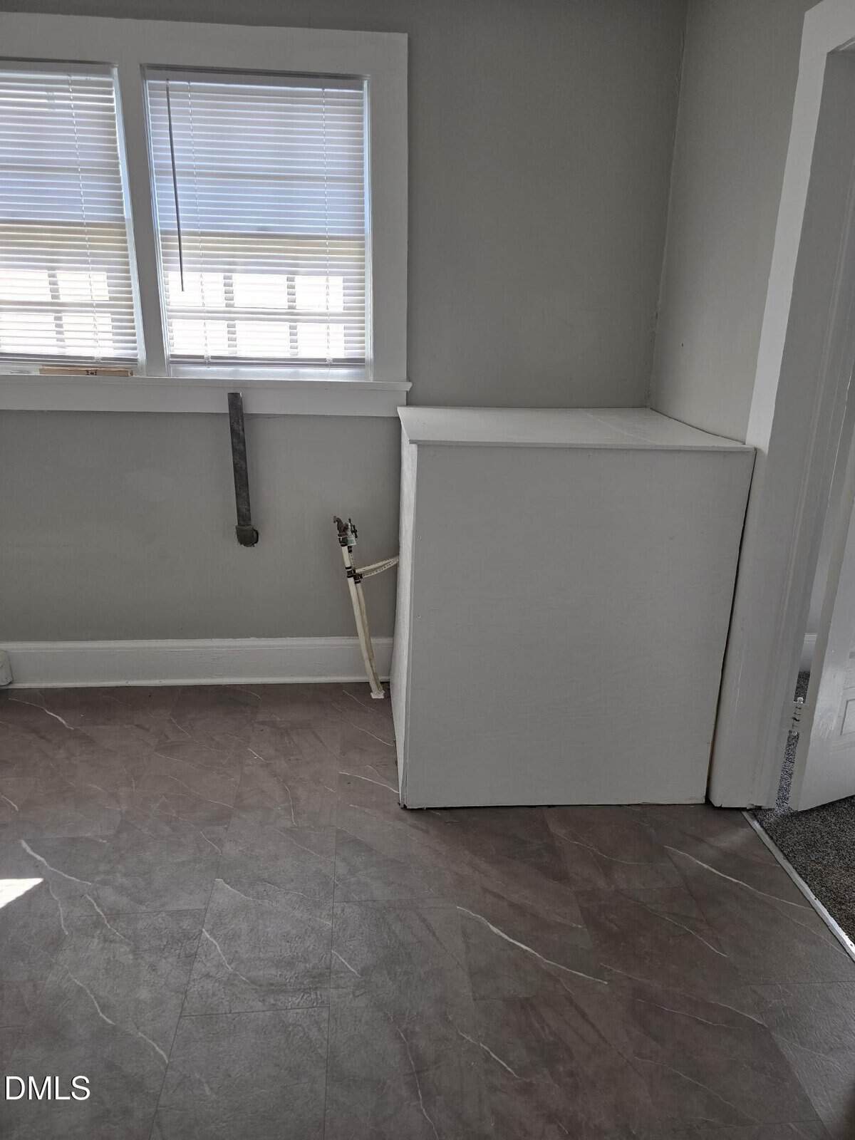 A small, empty laundry area with vinyl flooring, a covered utility hookup, exposed water pipes, and two windows with blinds letting in natural light.