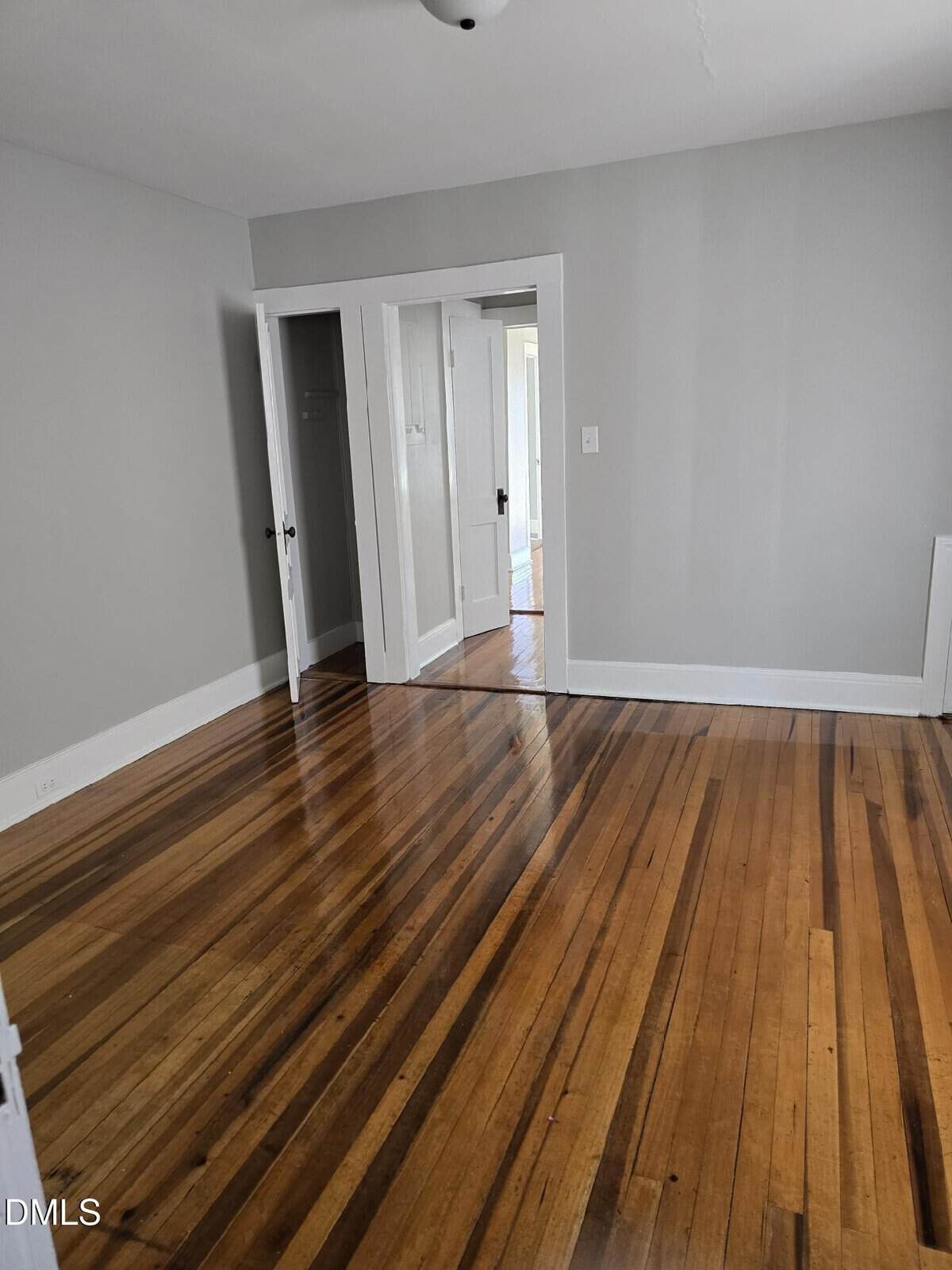 A bright, empty room with light gray walls, white trim, and polished hardwood floors. There are two doorways, one with a closed white door and another leading to a lit hallway.