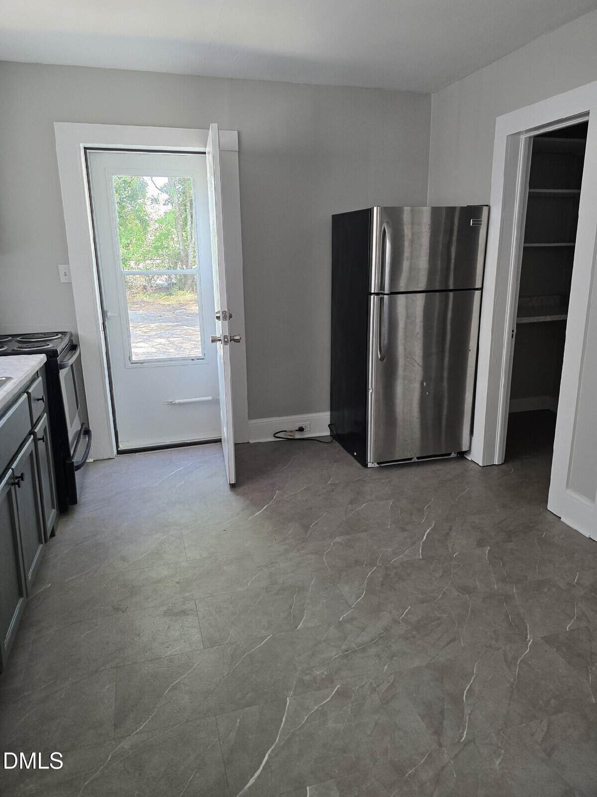 A kitchen with gray cabinets, a stainless steel refrigerator, a stove, and an open door leading outside. There is also an open pantry to the right of the fridge.