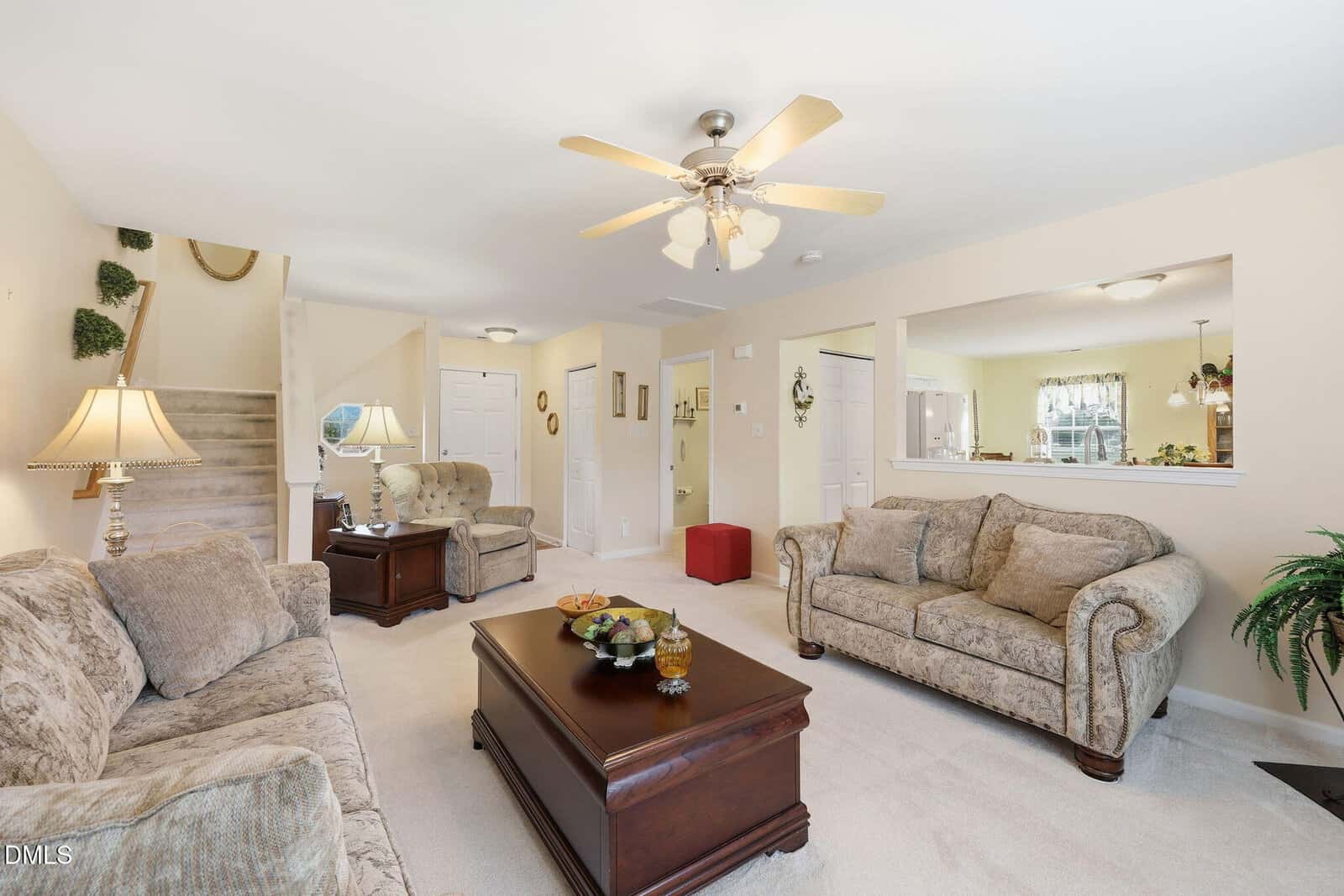 Bright living room with two beige sofas, matching armchair, coffee table, ceiling fan, lamps, and open view to the kitchen. Stairs and hallway are visible; decor is neutral with plants and framed pictures.