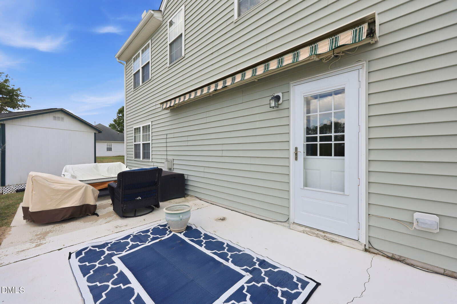 A backyard patio with a white door, green siding, an awning, outdoor furniture, a blue patterned rug, a small planter, and a shed in the background under a blue sky.