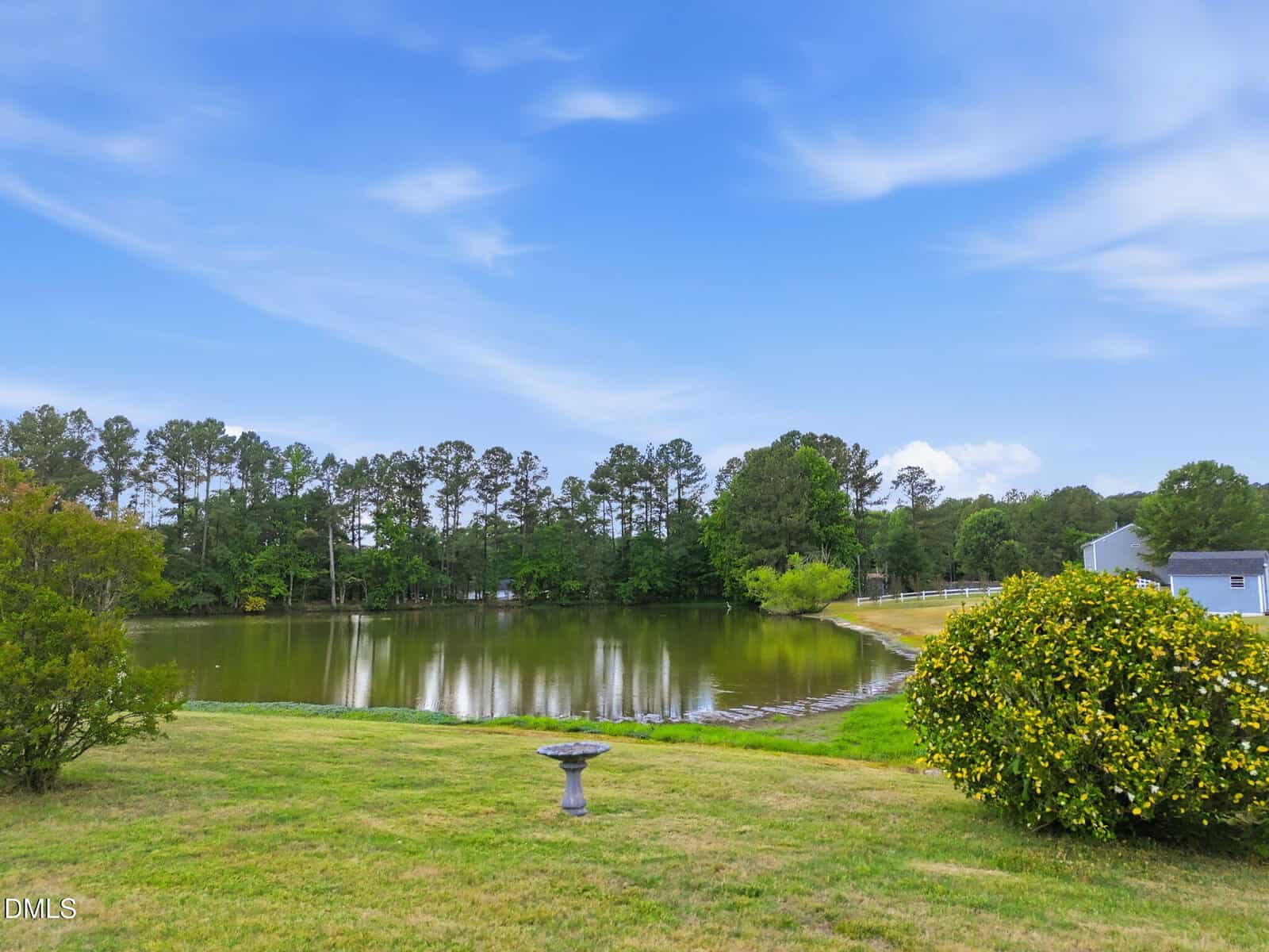 A grassy yard with a birdbath overlooks a small pond surrounded by trees and a partly cloudy blue sky. A few houses and bushes are visible on the right side of the image.