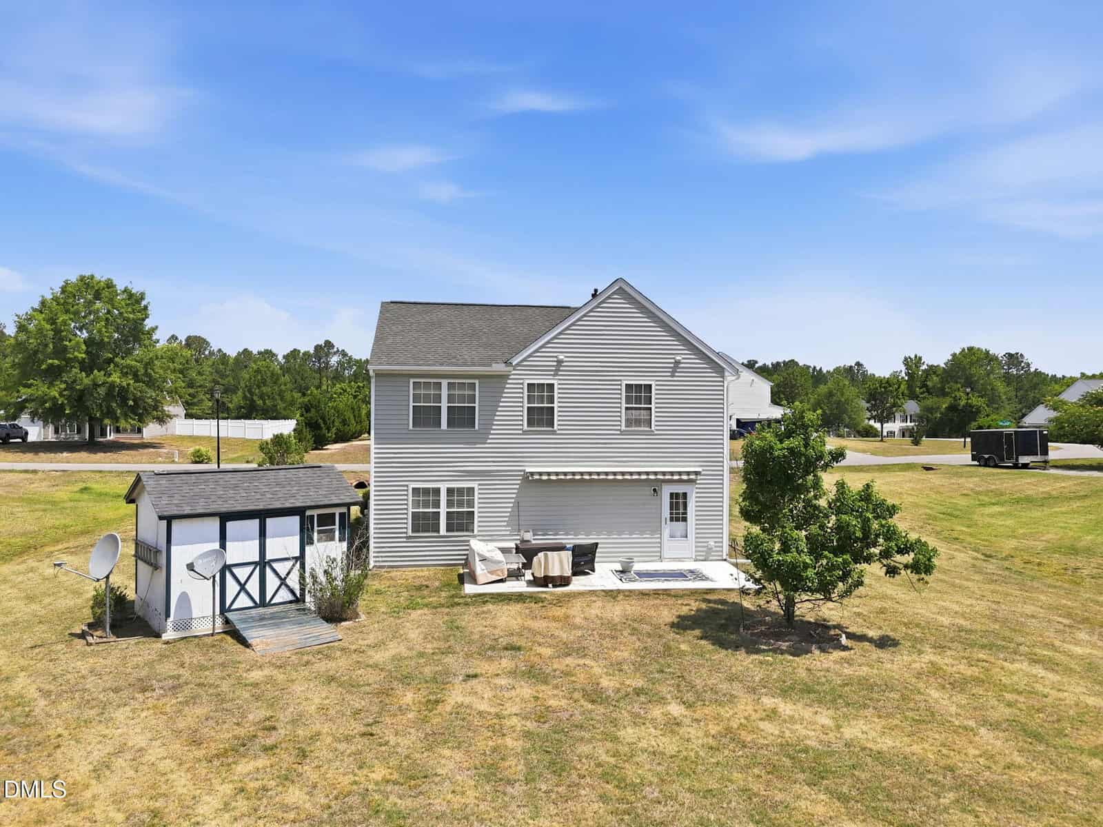 Two-story gray house with white trim and an attached patio set, next to a small blue and white shed. The house is surrounded by a large, grassy yard with a few trees, under a blue sky with light clouds.