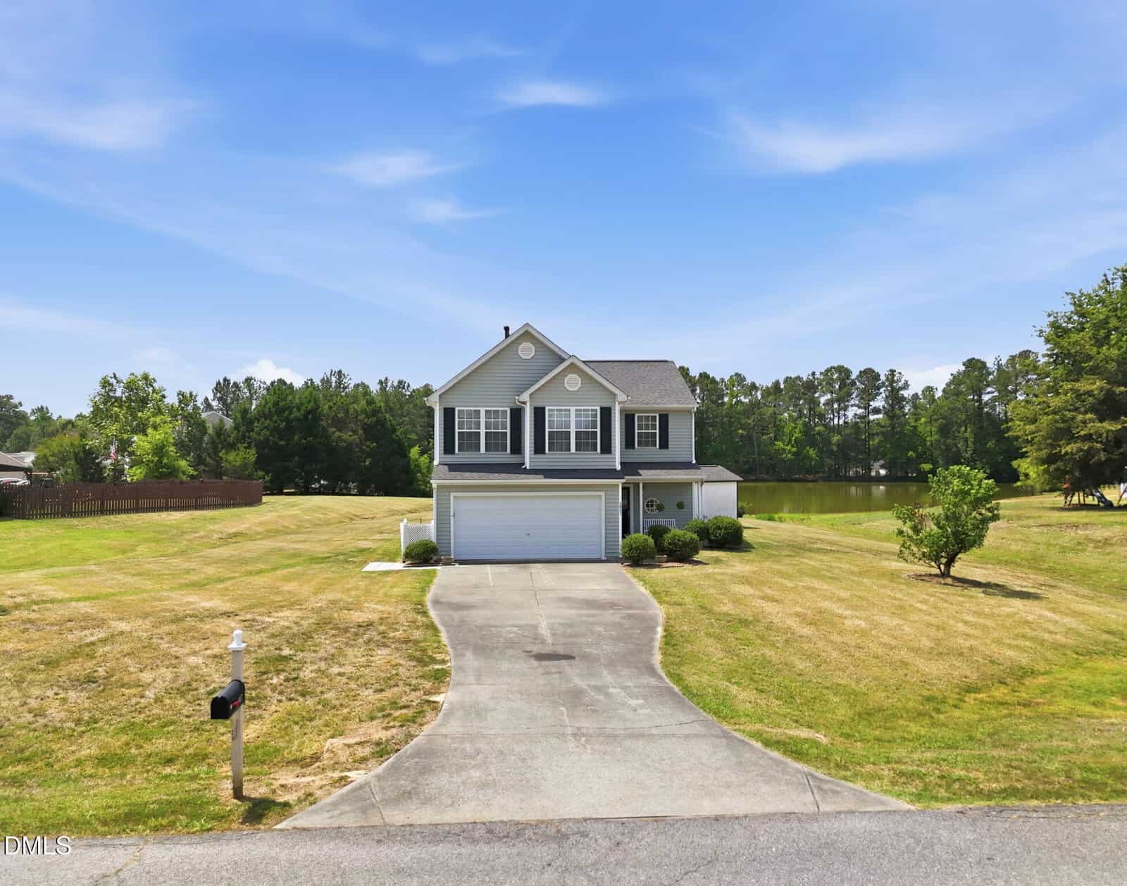 A two-story gray house with white trim and a double garage sits on a large, neatly mowed lawn at 2057 Hidden Pond Drive in Creedmoor, featuring a curved driveway, surrounded by trees and a lake visible under a blue sky.