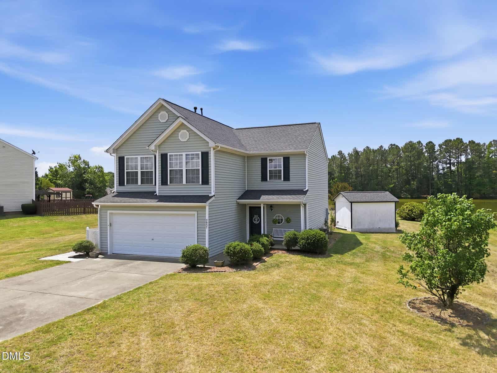 A two-story gray house with white trim and a double garage sits on a large, well-kept lawn. There is a small shed in the backyard, a tree to the right, and trees in the distance under a blue sky.