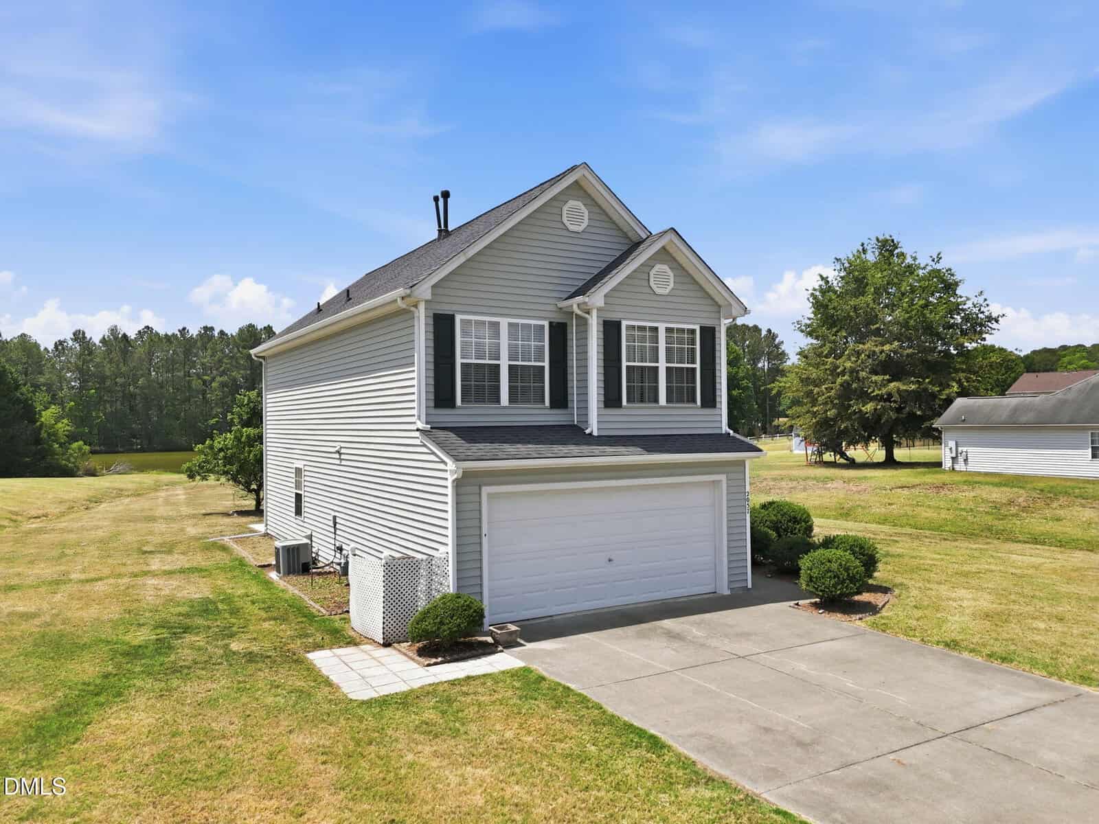 Two-story suburban house with gray siding and white trim, double garage, and small bushes along the driveway. The house is surrounded by a large grassy yard with trees in the background under a blue sky.