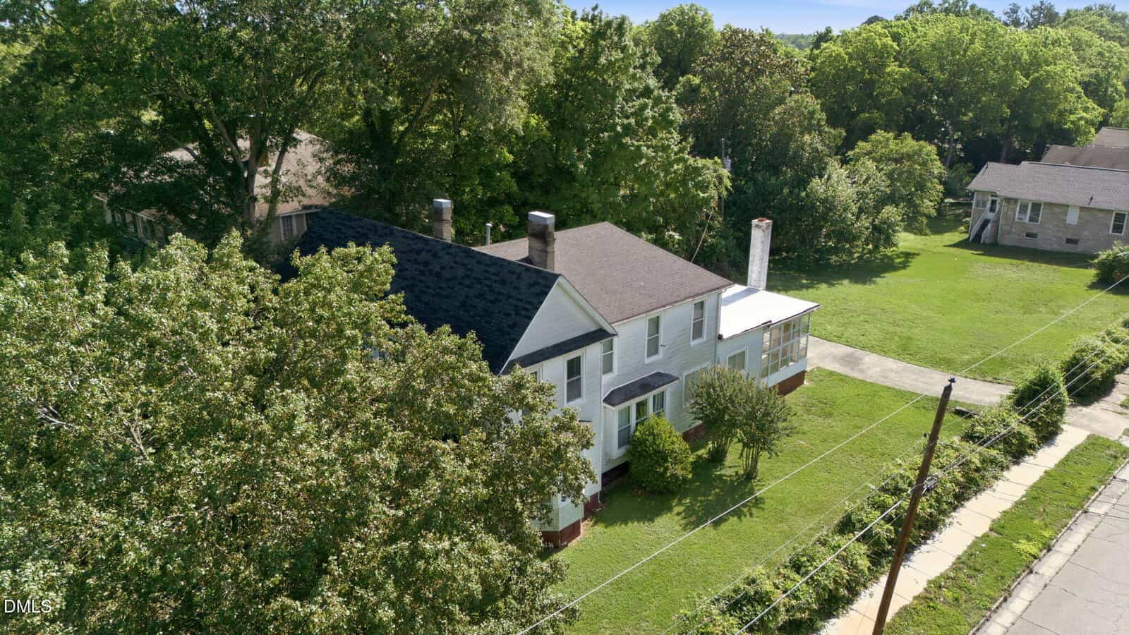 Aerial view of a two-story white house with a dark roof, surrounded by trees and a green lawn. A driveway runs alongside the house, and another house is visible in the background.